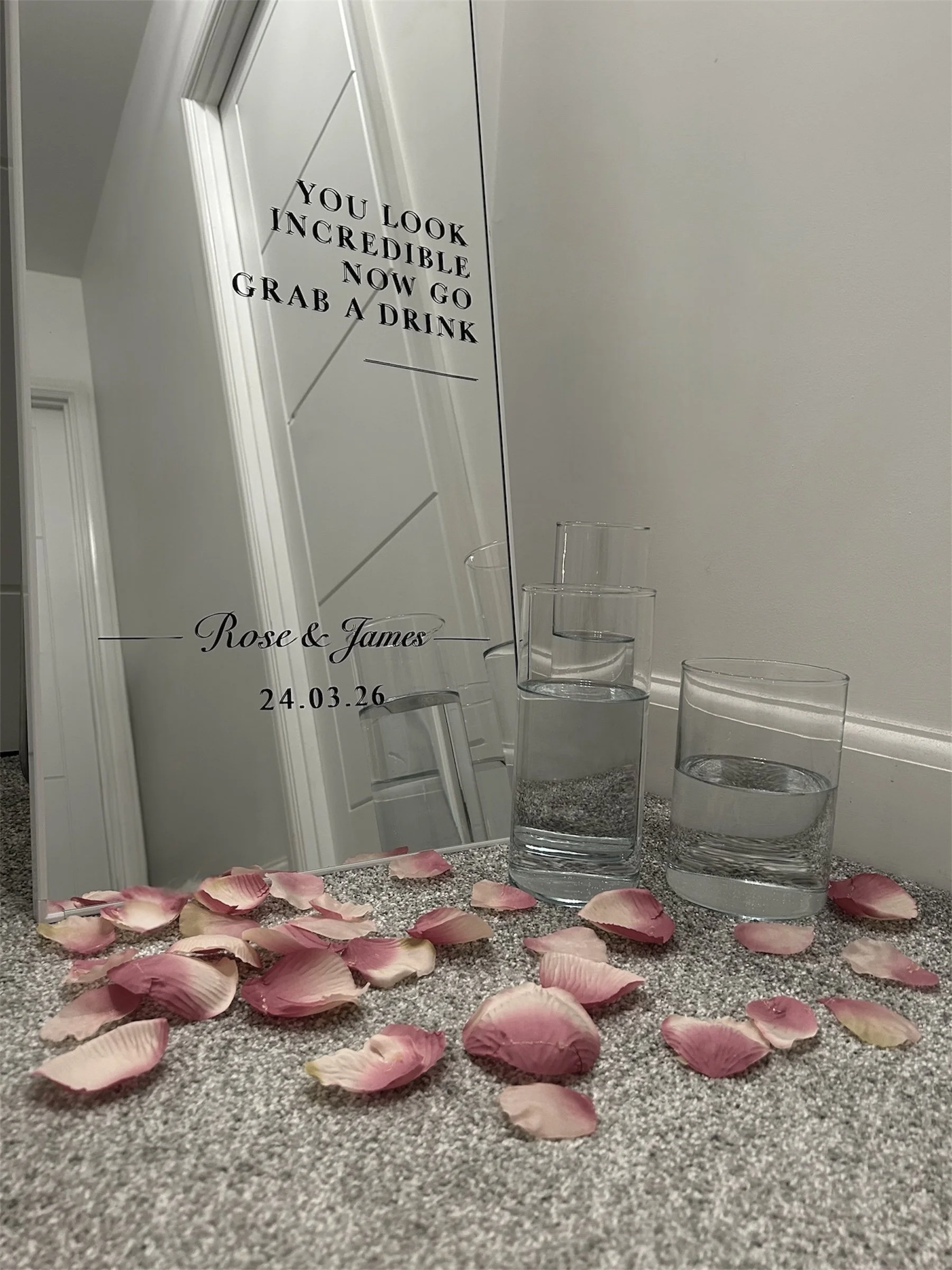 A mirror with an engraved message, two glasses of water, and pink rose petals scattered on a carpeted floor.