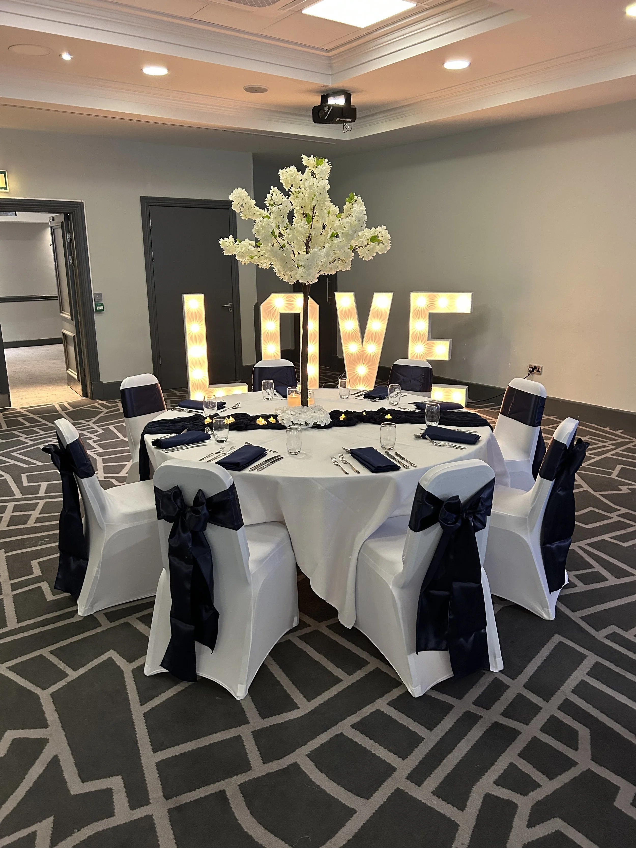 A decorated banquet table with white tablecloth, black napkins, and glasses, surrounded by white chairs with black sashes. A large illuminated sign spelling 'LOVE' with a white floral centerpiece resembling a tree is in the background.