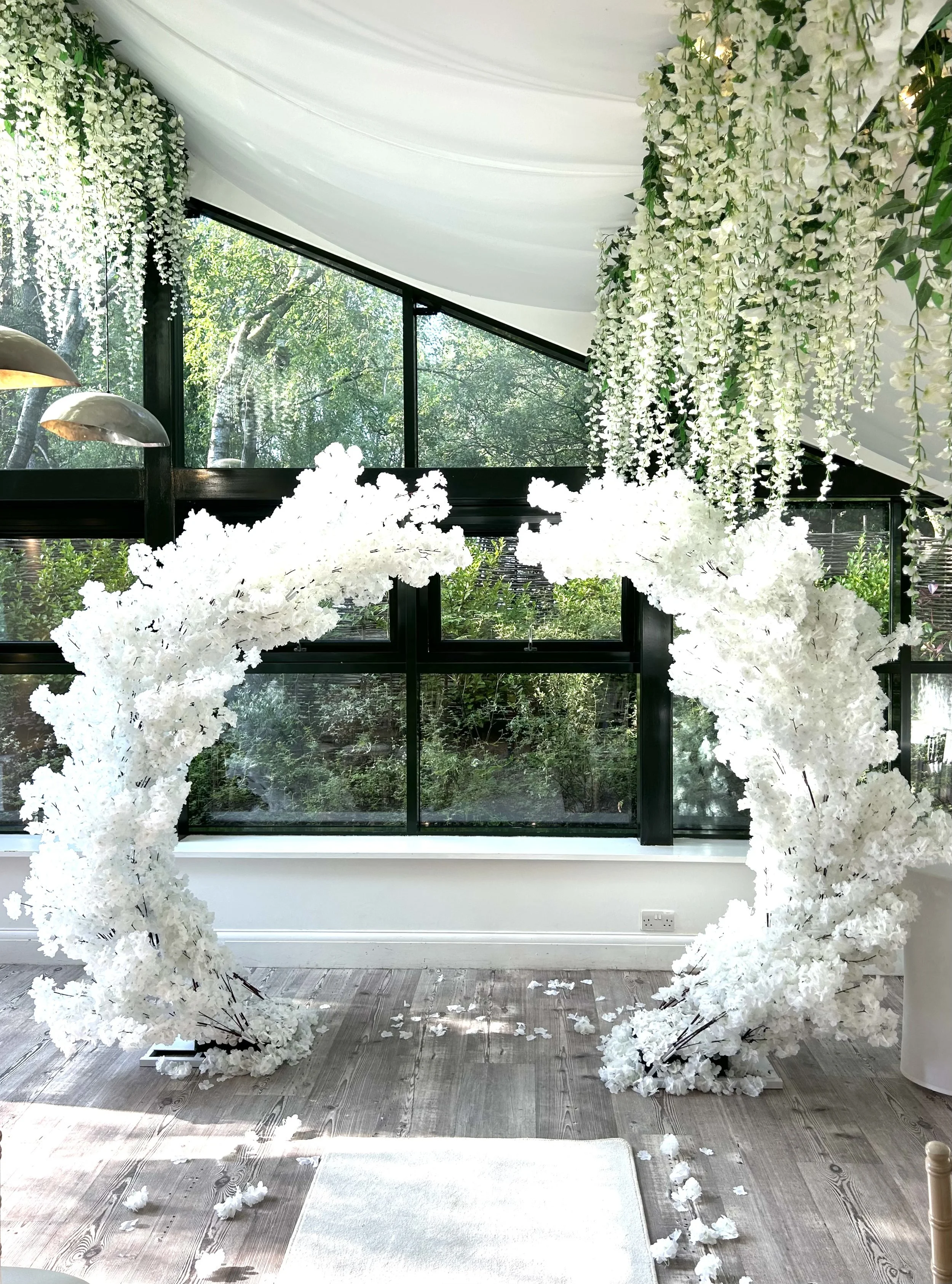 Indoor floral decoration with white flowers forming a semi-circle, hanging white flowers from the ceiling, and a glass window showing greenery outside.