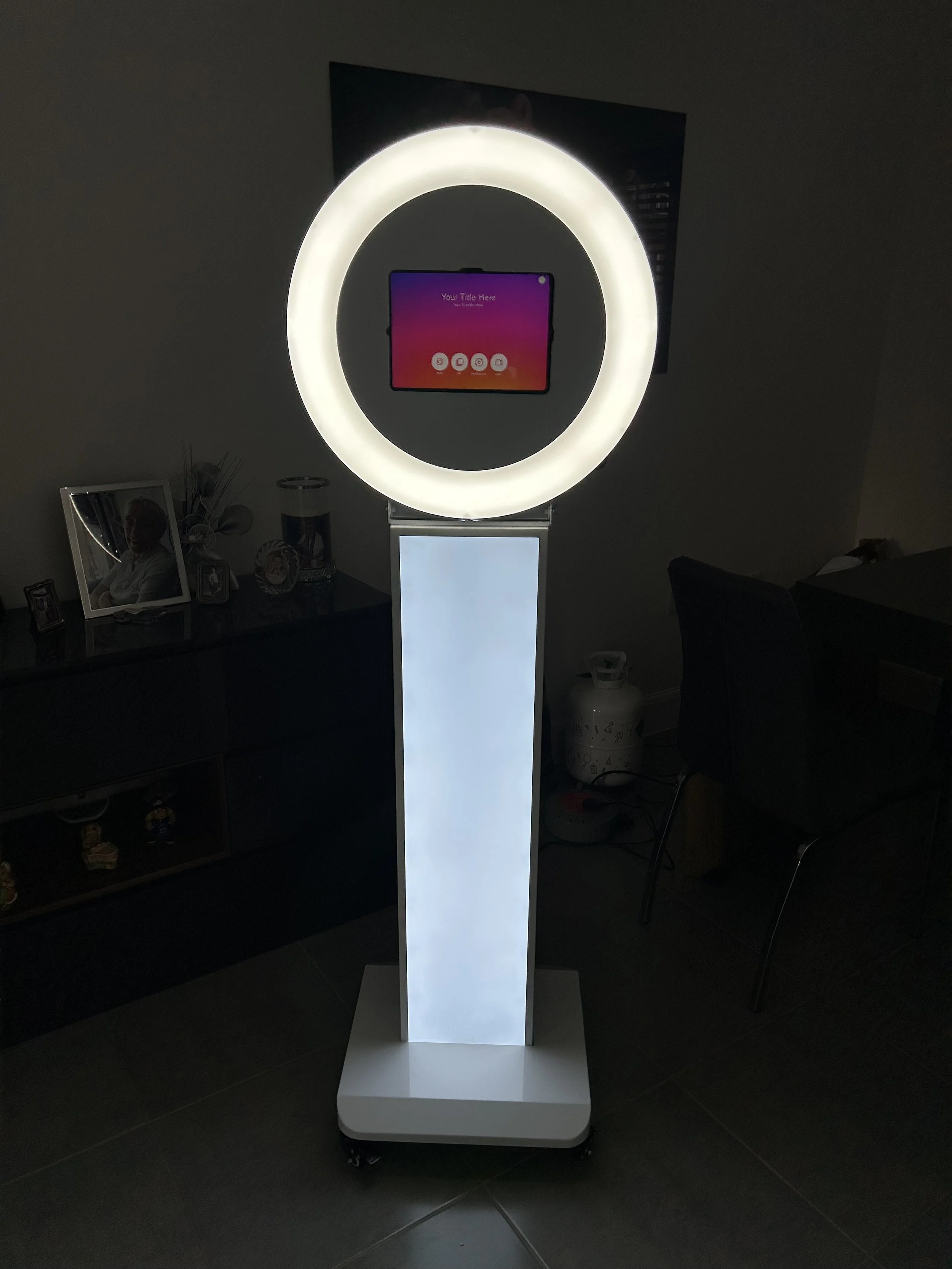 Photo booth with illuminated ring light, tablet in the center, on a white stand with wheels, set in a dark room.