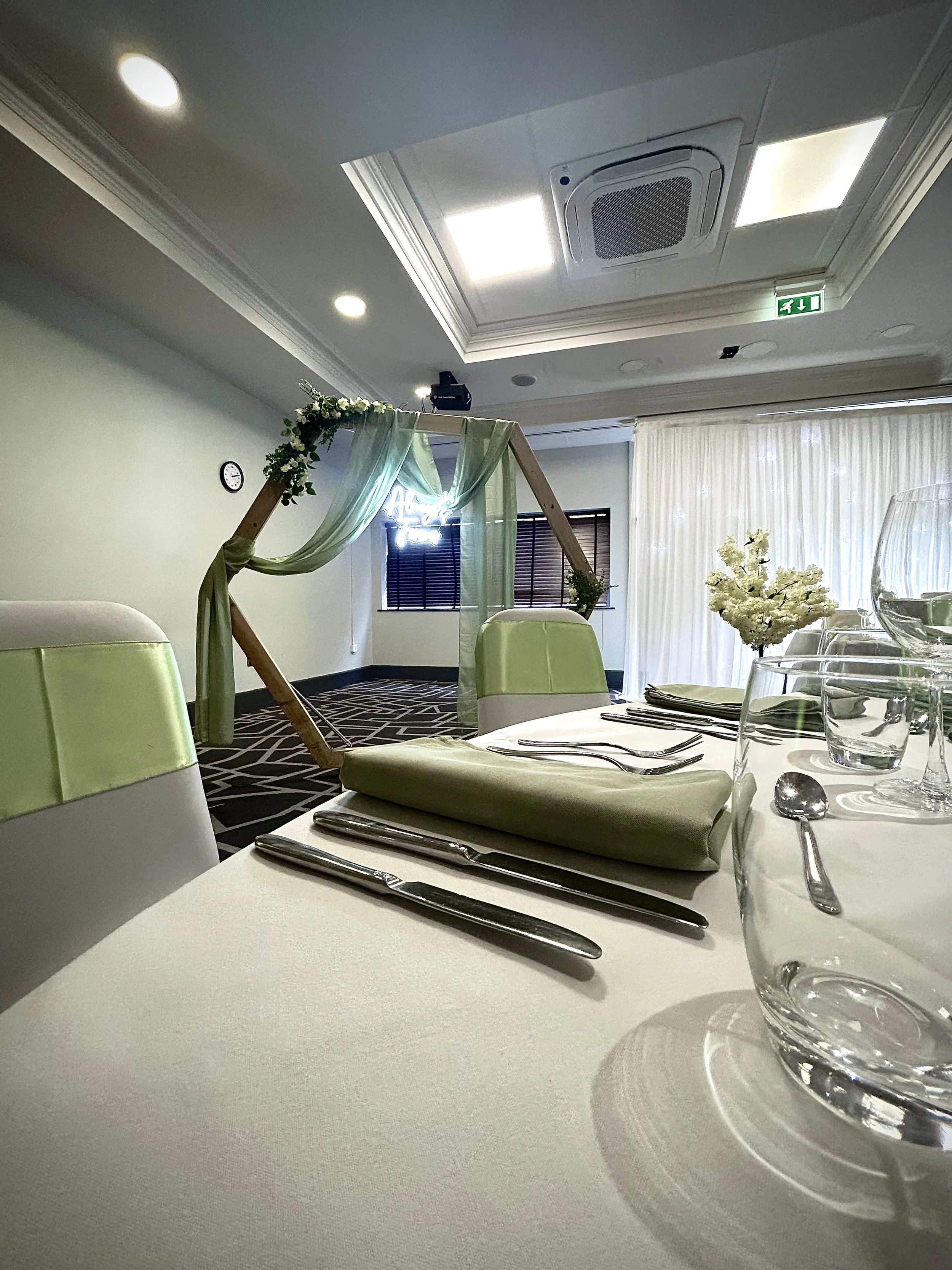 Wedding reception table with green and white chairs, set with silverware, glasses, and napkins. In the background, a wooden arch with sheer green fabric and flowers.