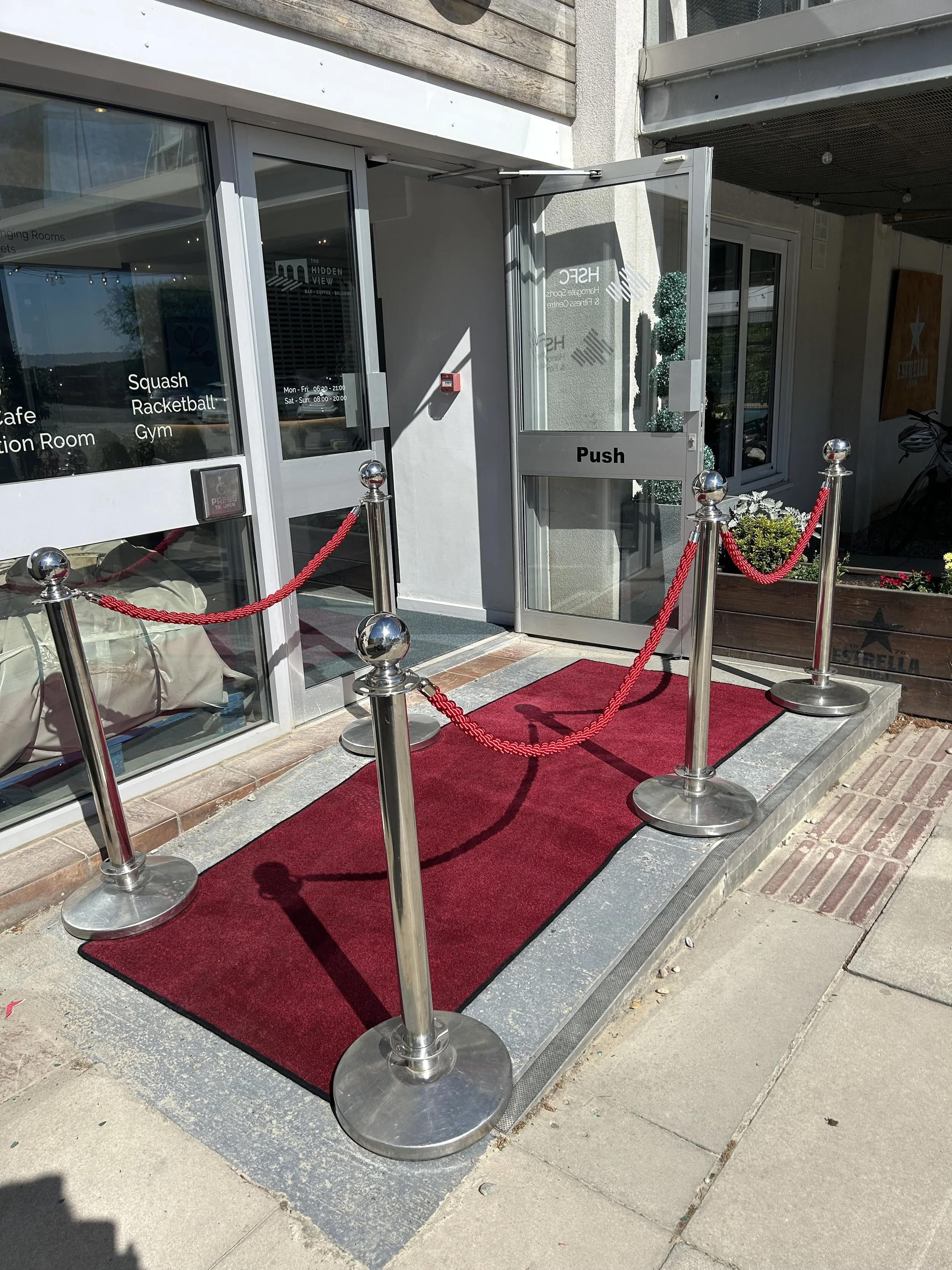 Red carpet entryway with silver stanchions and red velvet ropes leading to a glass door marked 'Push' at a modern building entrance.
