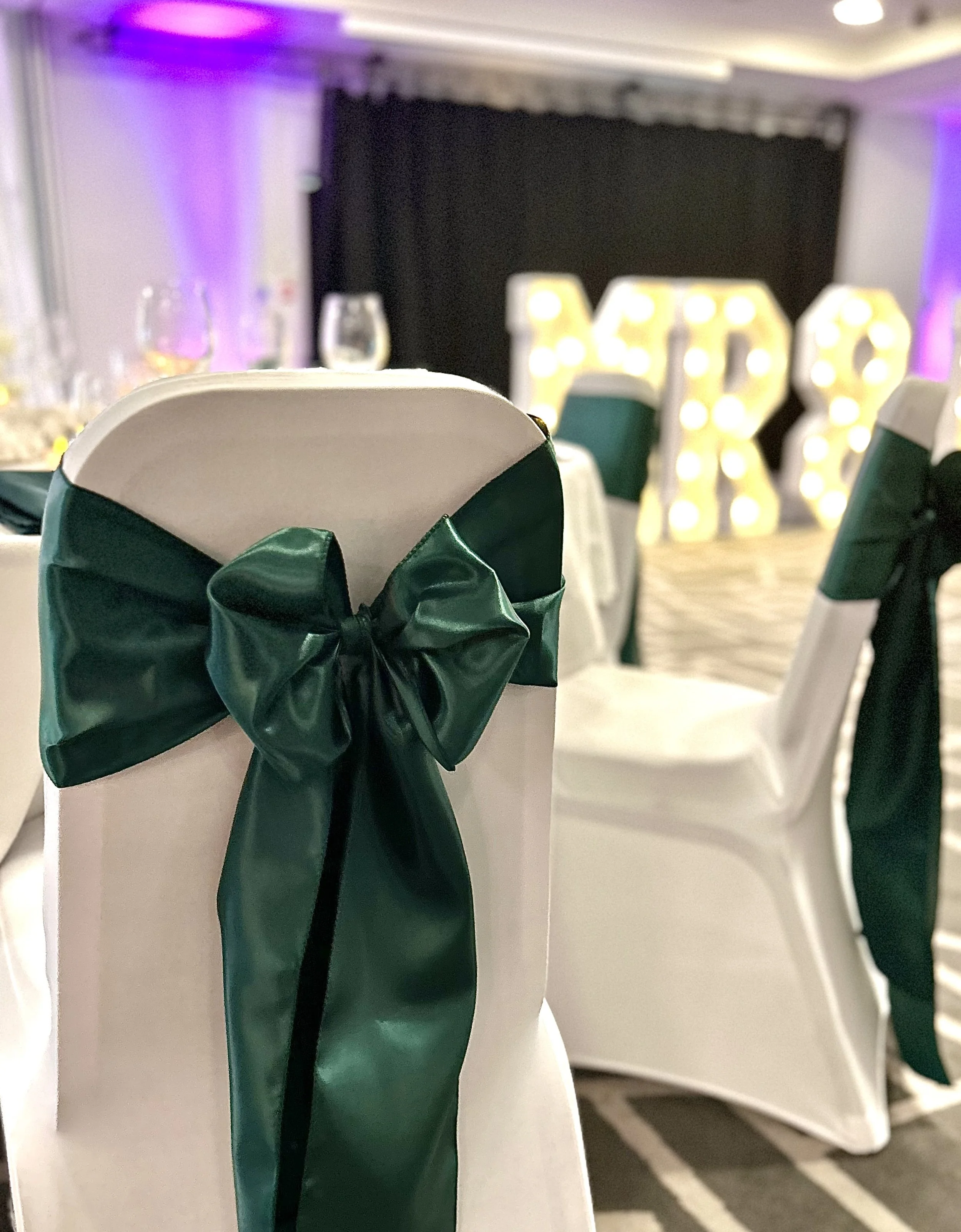Wedding reception table setting with white chairs adorned with green satin sashes tied in bows. In the background, illuminated marquee letters spell out "PERS".