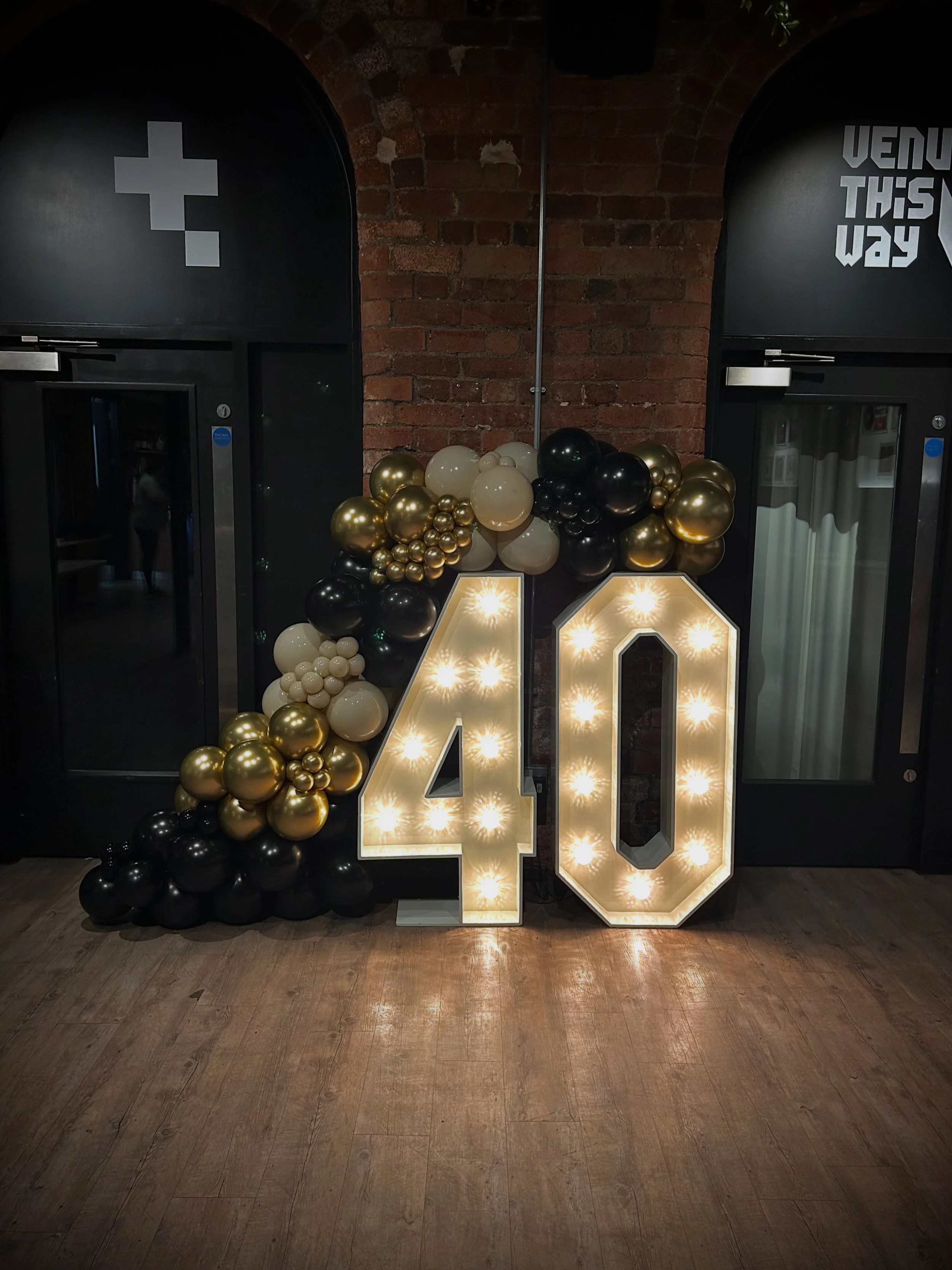 Decorative display featuring large illuminated number 40, surrounded by black, white, and gold balloons, set against a brick wall in an indoor venue.