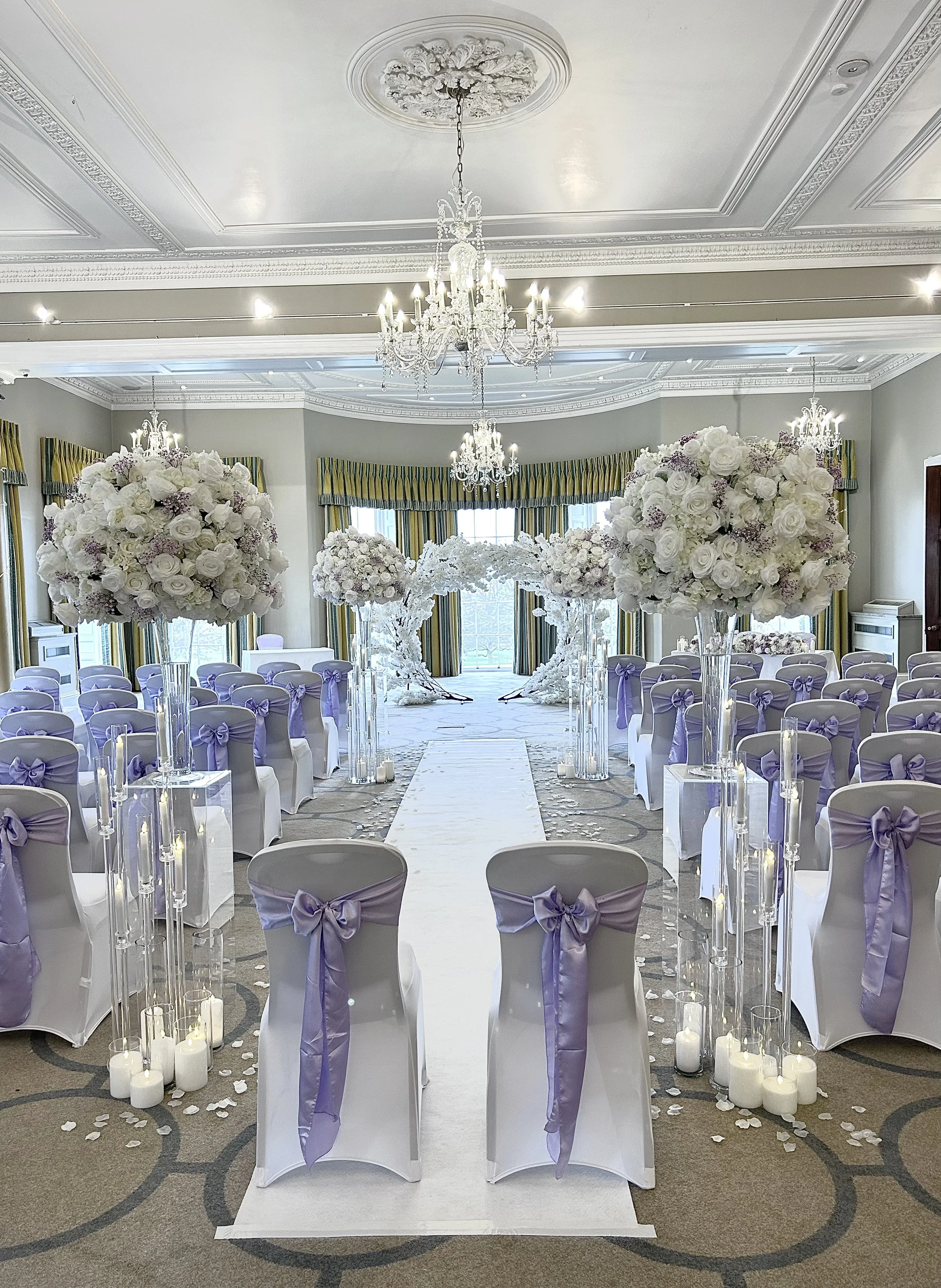 Elegant wedding ceremony setup with white chairs decorated with purple bows, large floral arrangements in tall glass vases, white flower petals scattered on the aisle, and a backdrop of a white decorated arch in a bright room with chandeliers and gold and green curtains.