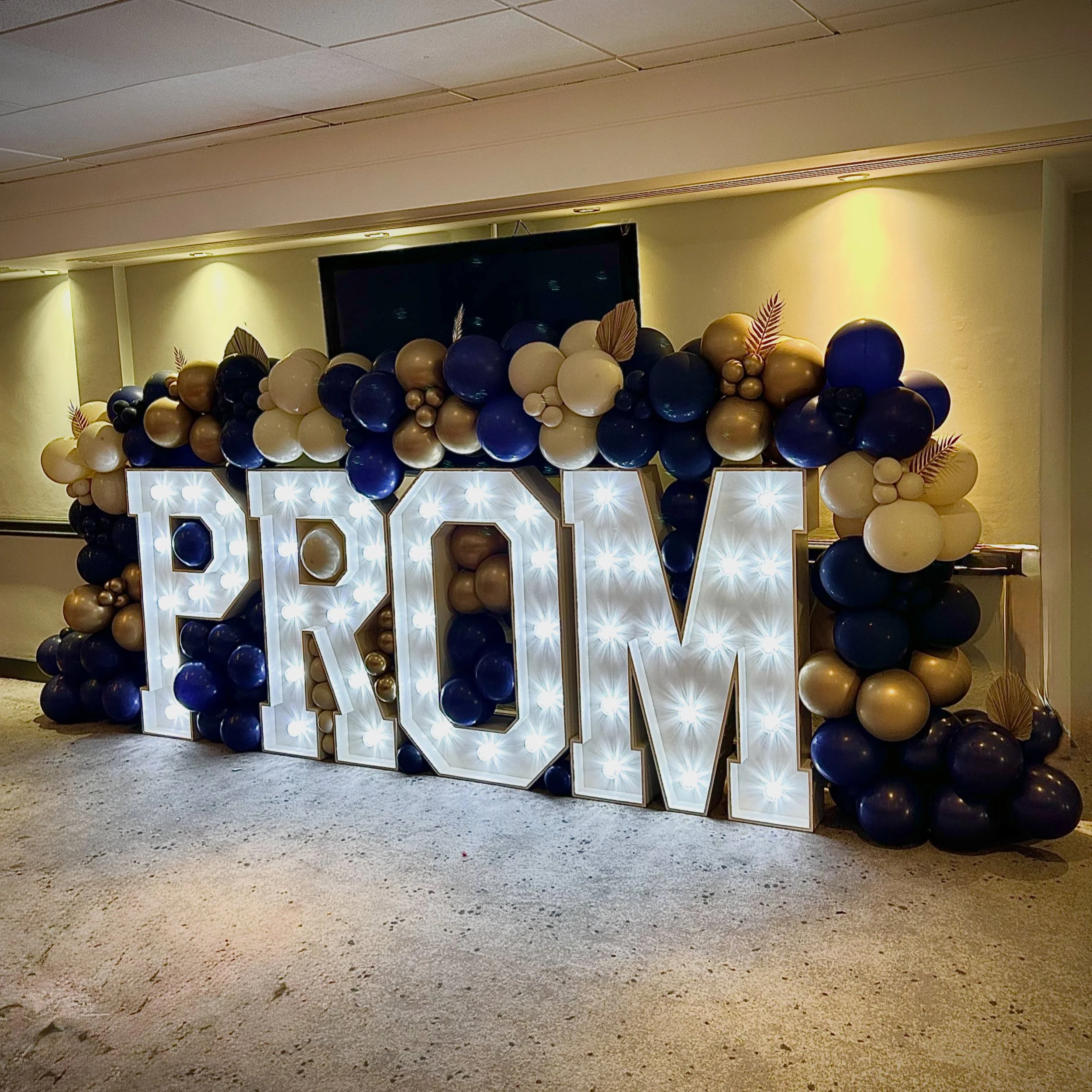 Decorative balloon arch with blue, white, gold, and black balloons and illuminated letter 'V' at an event.