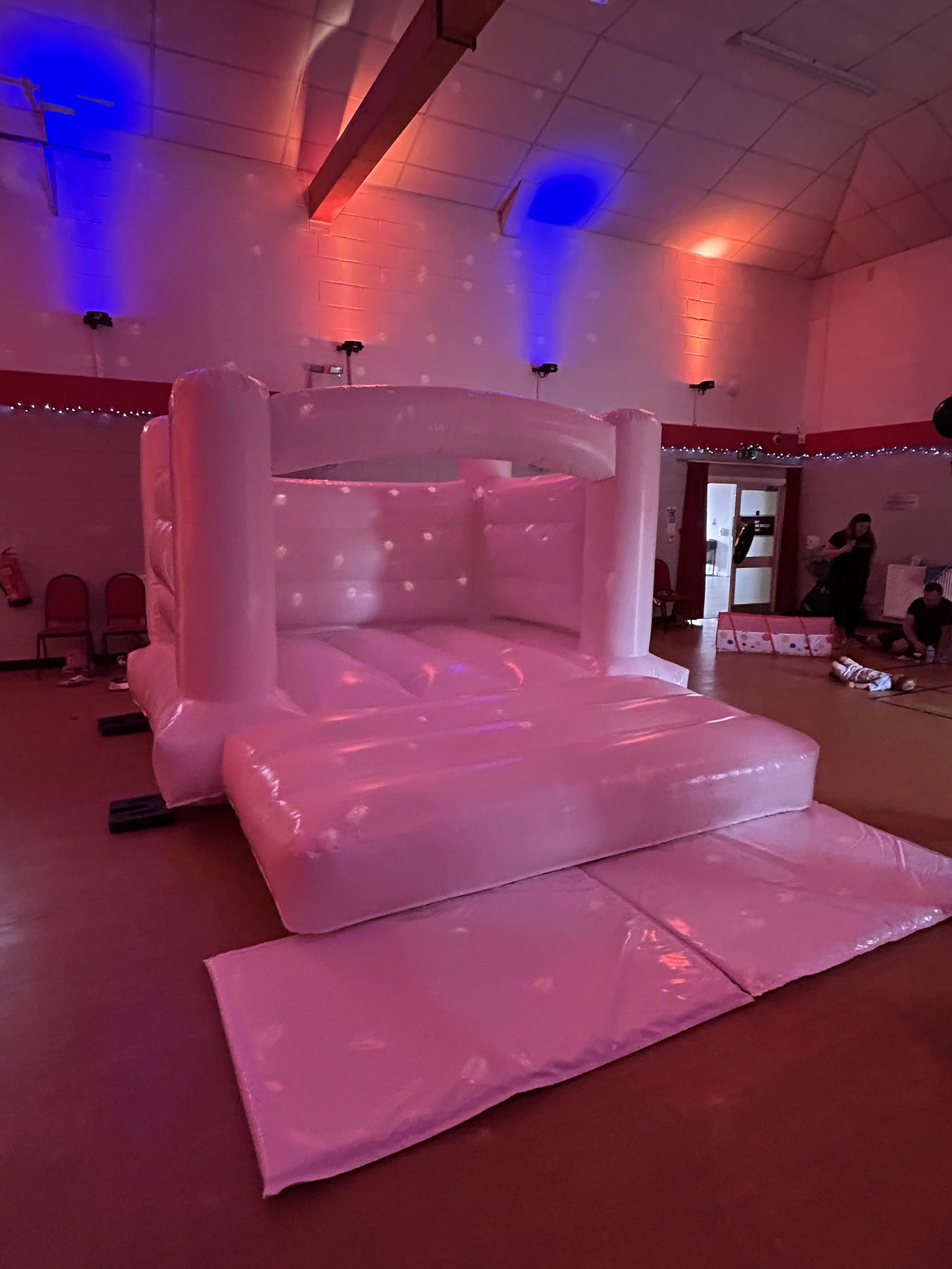 Inflatable pink castle with a slide in a large indoor hall, decorated with colorful lighting and people in the background.