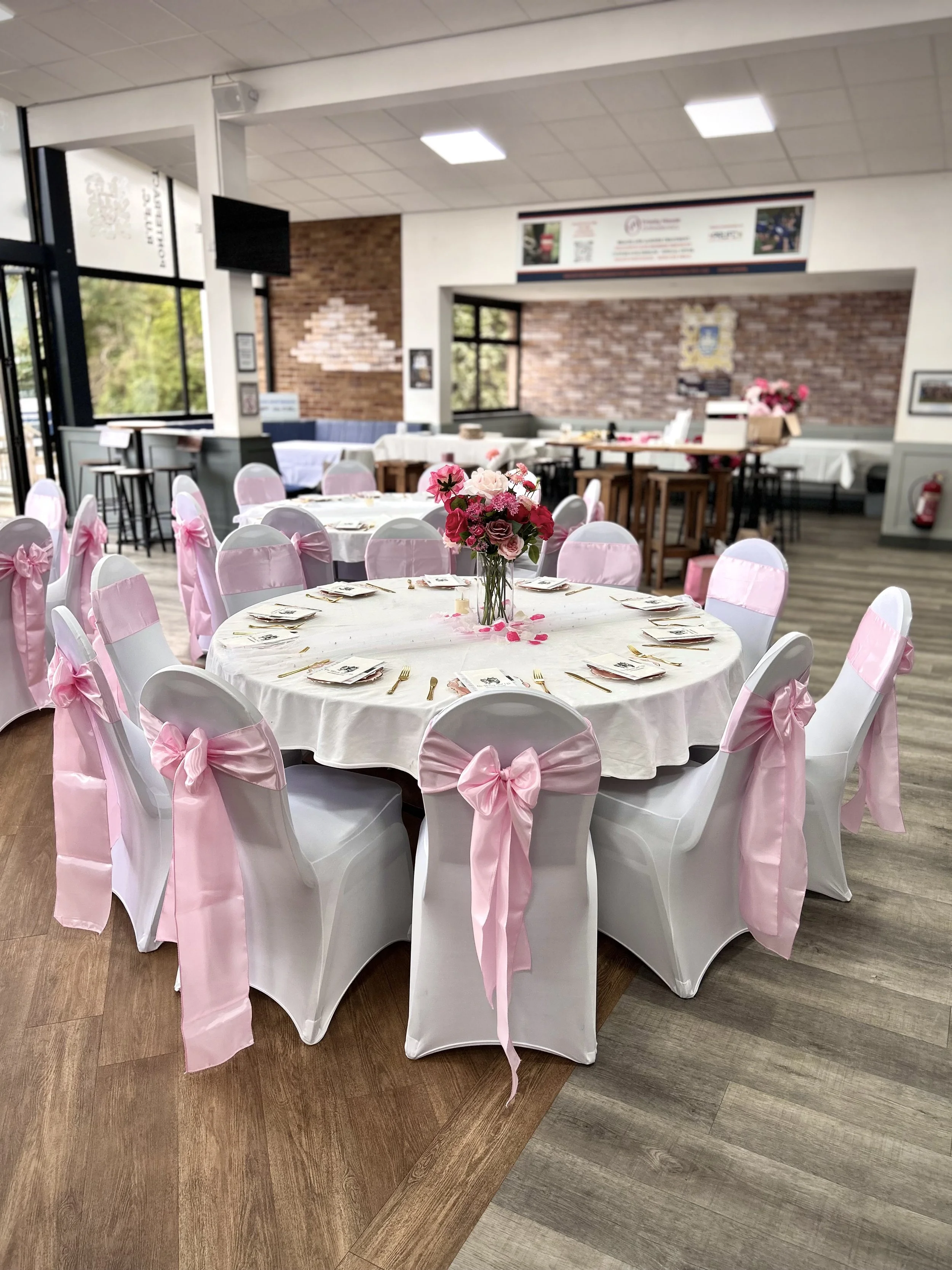 A decorated banquet hall with a round table covered with a white tablecloth, adorned with a pink floral centerpiece, surrounded by white chairs with pink sashes. The hall has brick and white walls, large windows, and a wooden floor transitioning into