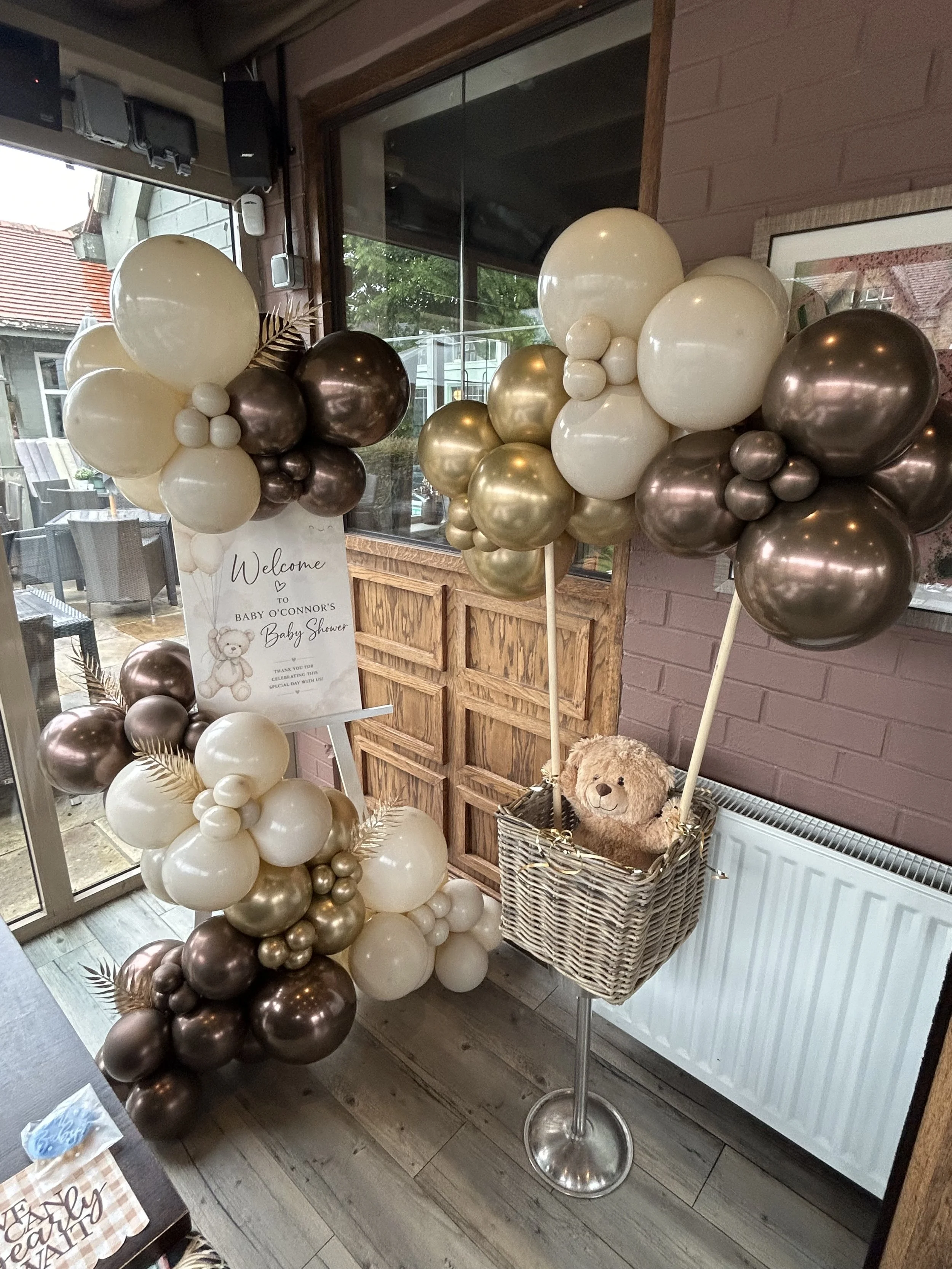 Balloon arrangement in shades of white, gold, and brown near a window at a baby shower, with a welcome sign and a teddy bear decoration in a basket.