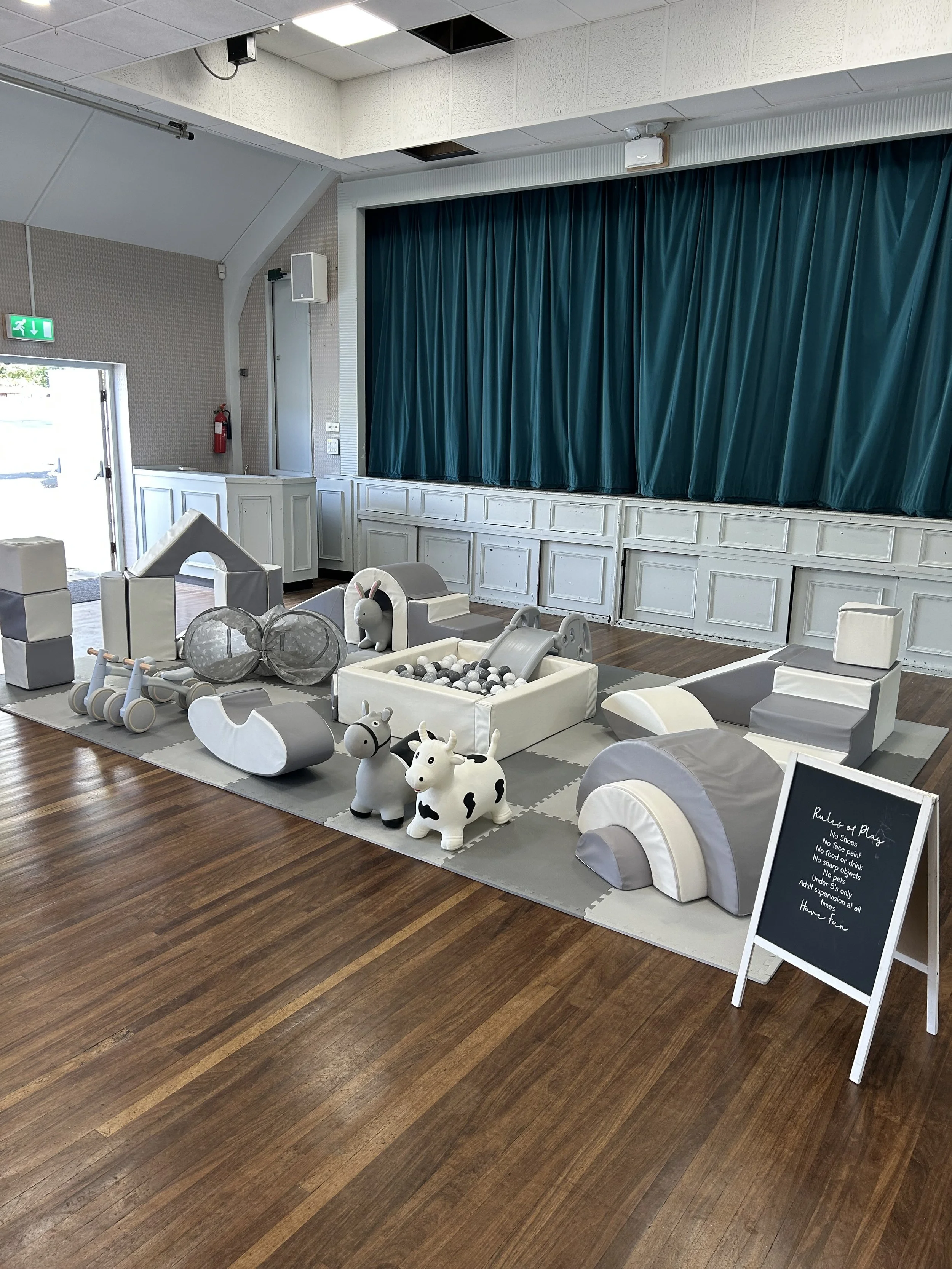 Empty indoor children's play area with soft grey and white toys, a small ball pit, stuffed animal toys, and a blue curtain backdrop.
