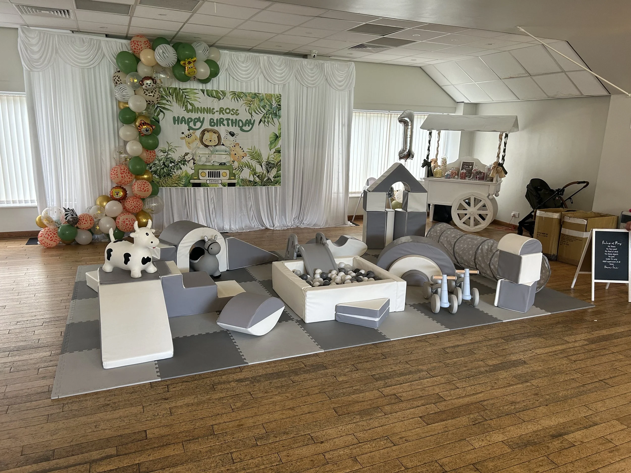 Indoor children's play area decorated for a birthday party with a jungle and animal theme. Features include soft foam climbing structures, a ball pit, animal-shaped toys, and a backdrop with balloons and a 