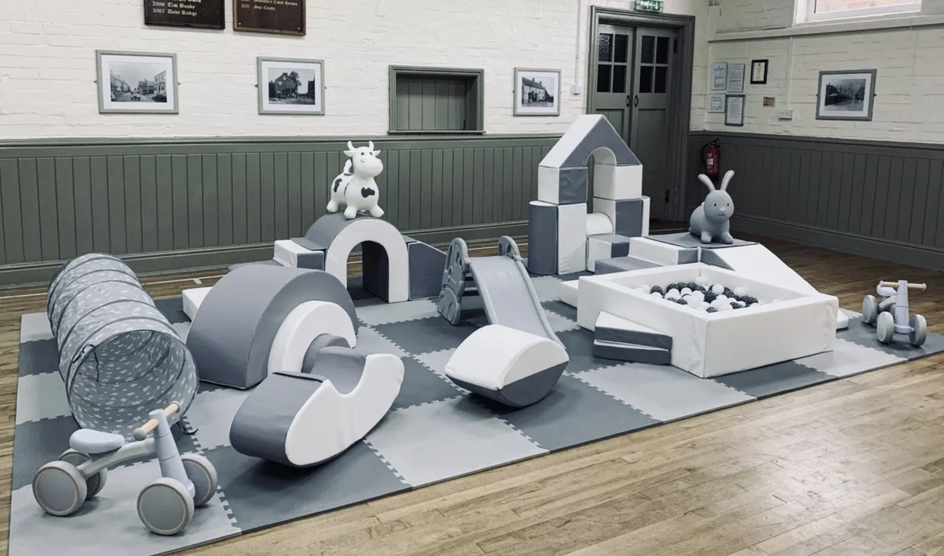 Indoor children's play area with soft gray and white toys and structures, including a tunnel, slide, animal figures, and a ball pit, on a gray mat with wooden flooring nearby.