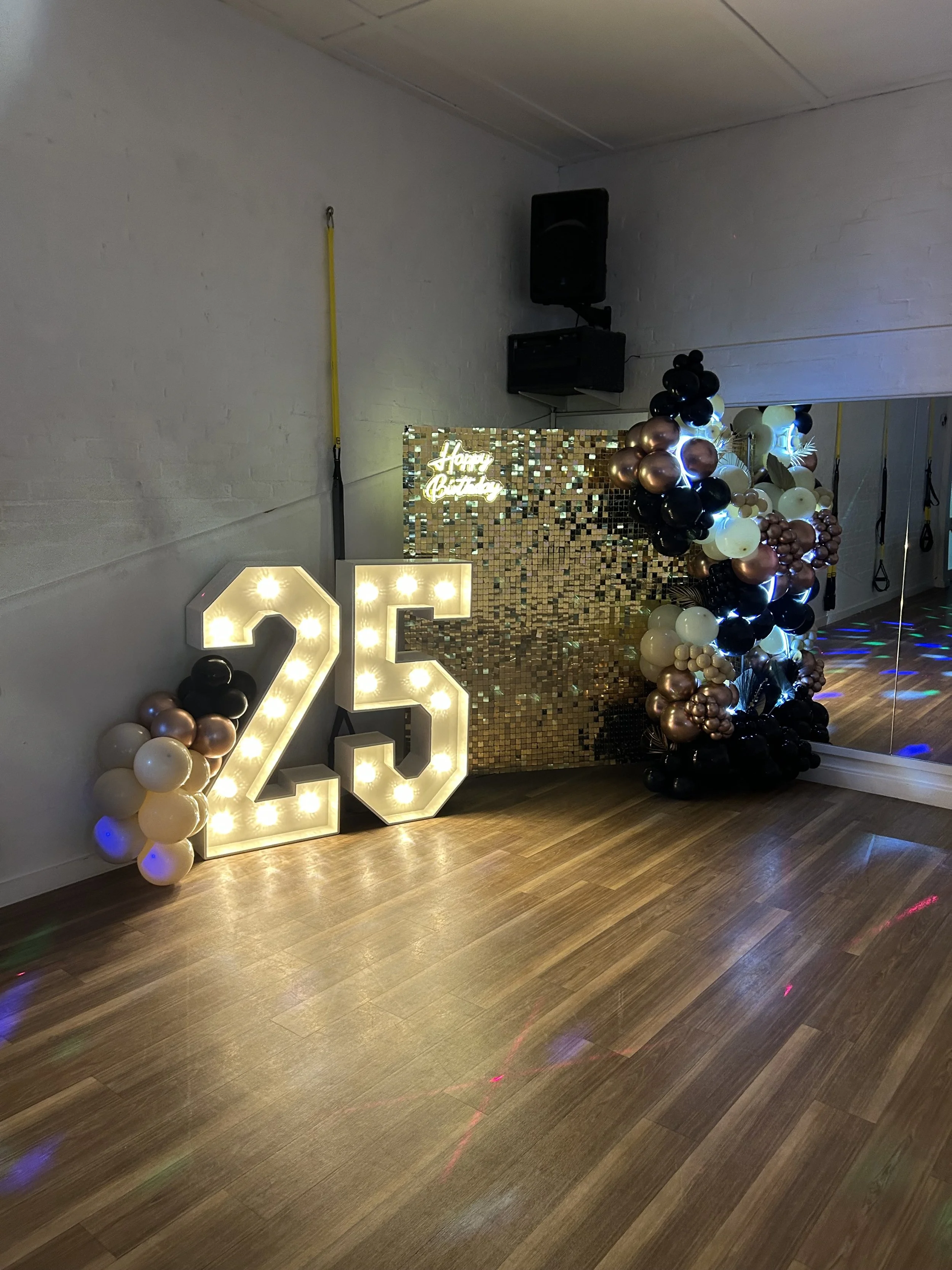 Decorations for a 25th birthday celebration include large illuminated number '25', a backdrop with small gold and black squares, balloon arrangements in black, gold, white, and rose gold, and a small neon sign that says 'Happy Birthday'