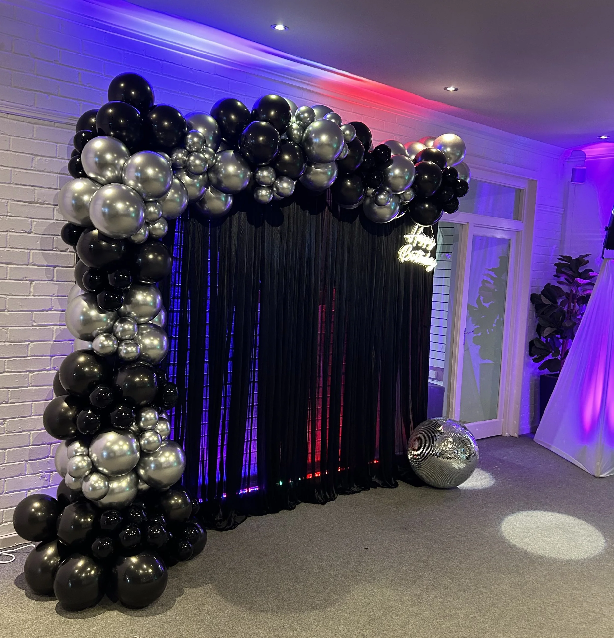 Black, silver, and dark gray balloon arch with black curtains and a disco ball in a decorated indoor party space with purple and pink lighting.