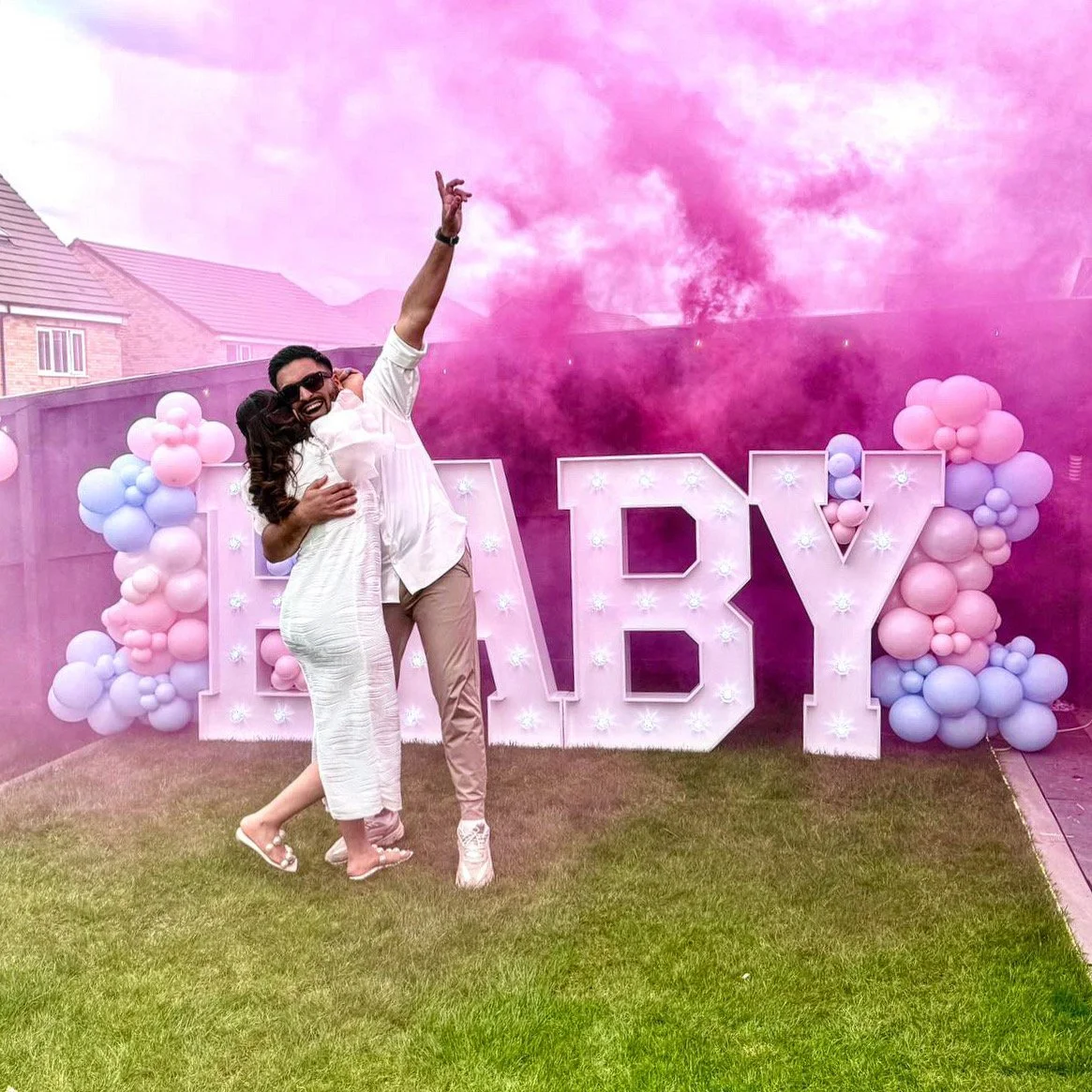 A couple celebrating at a backyard party with pink and purple balloons and a large marquee sign spelling 'BABY'. Pink smoke in the background.