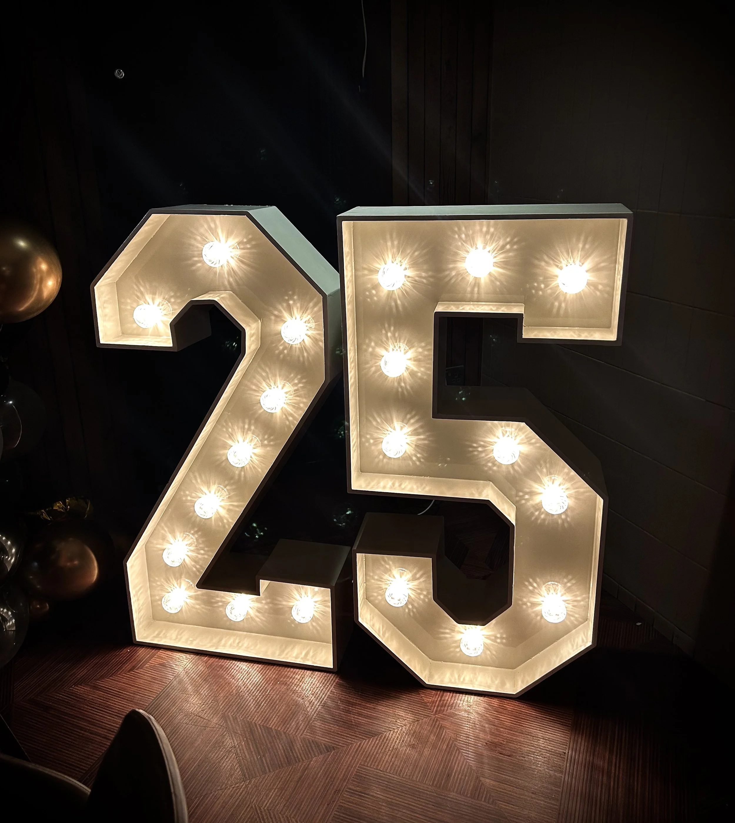 Large illuminated number '25' sign with light bulbs, placed on a wooden floor against a dark wall.
