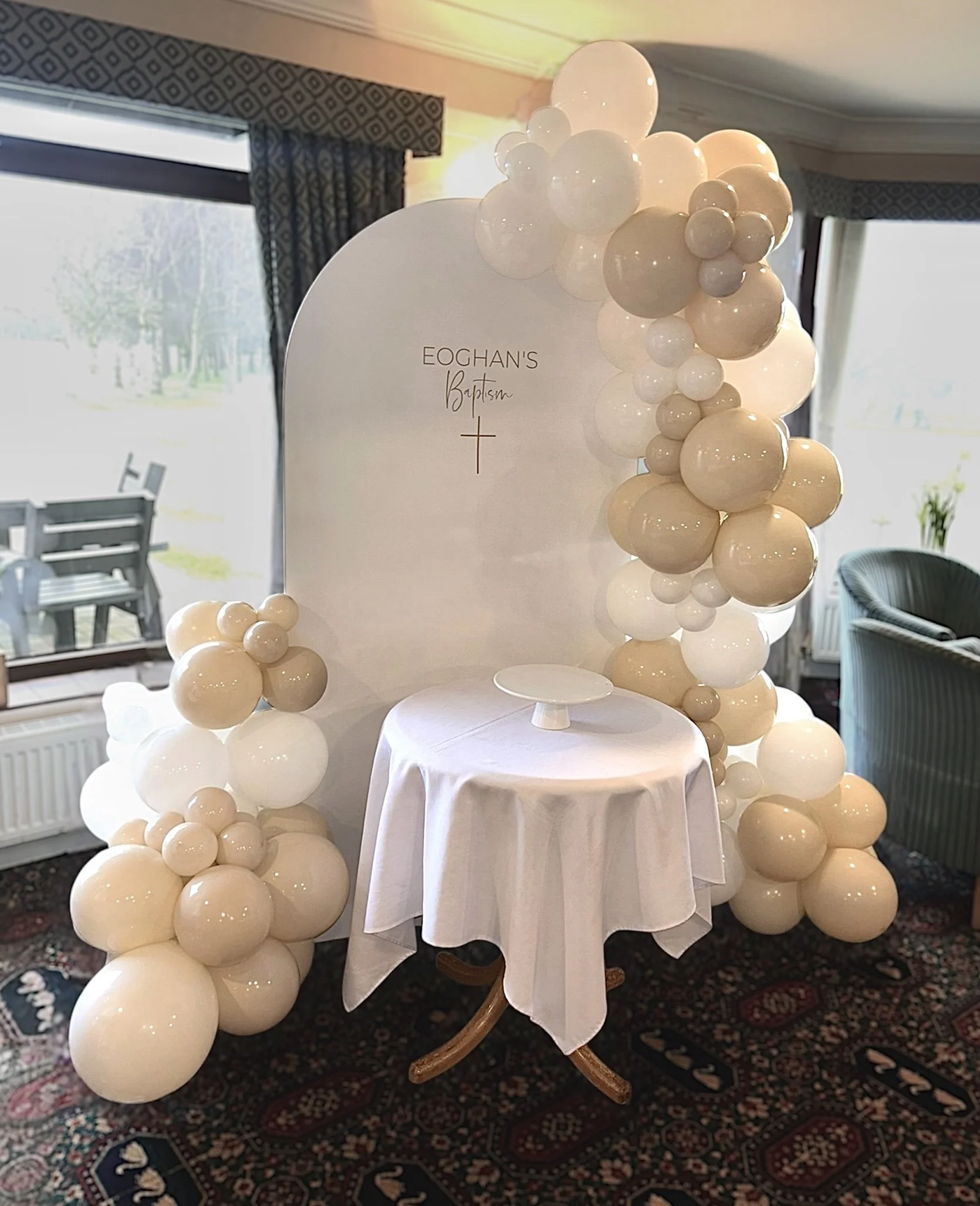 Decorative backdrop with balloons for a baptism celebration, featuring a white oval board with the text 'Eoghan's Baptism' and a cross, set in a room with windows and curtains.
