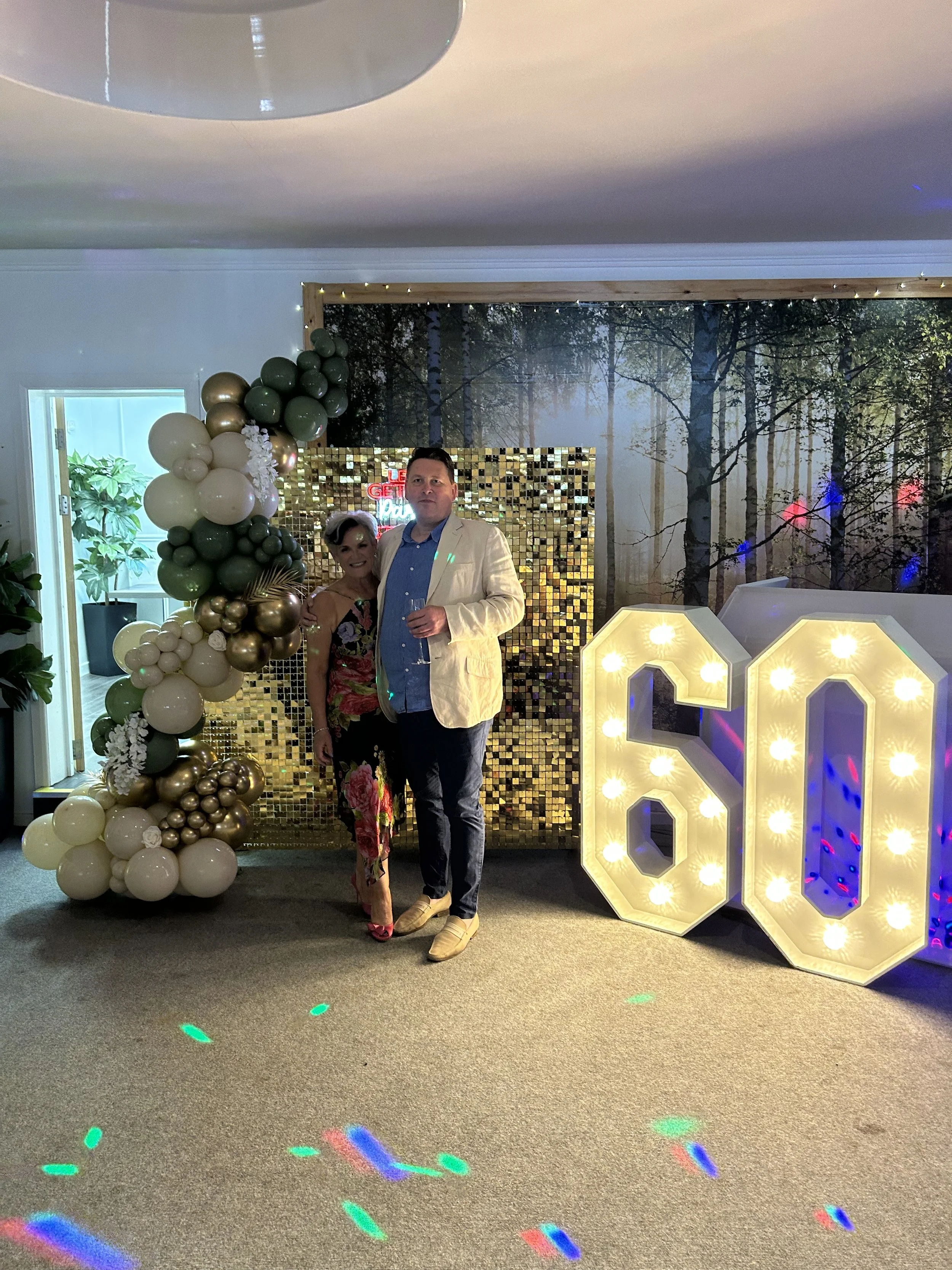 Two people standing together at a 60th birthday celebration with large illuminated number signs, a gold sequined backdrop, and balloon decorations.