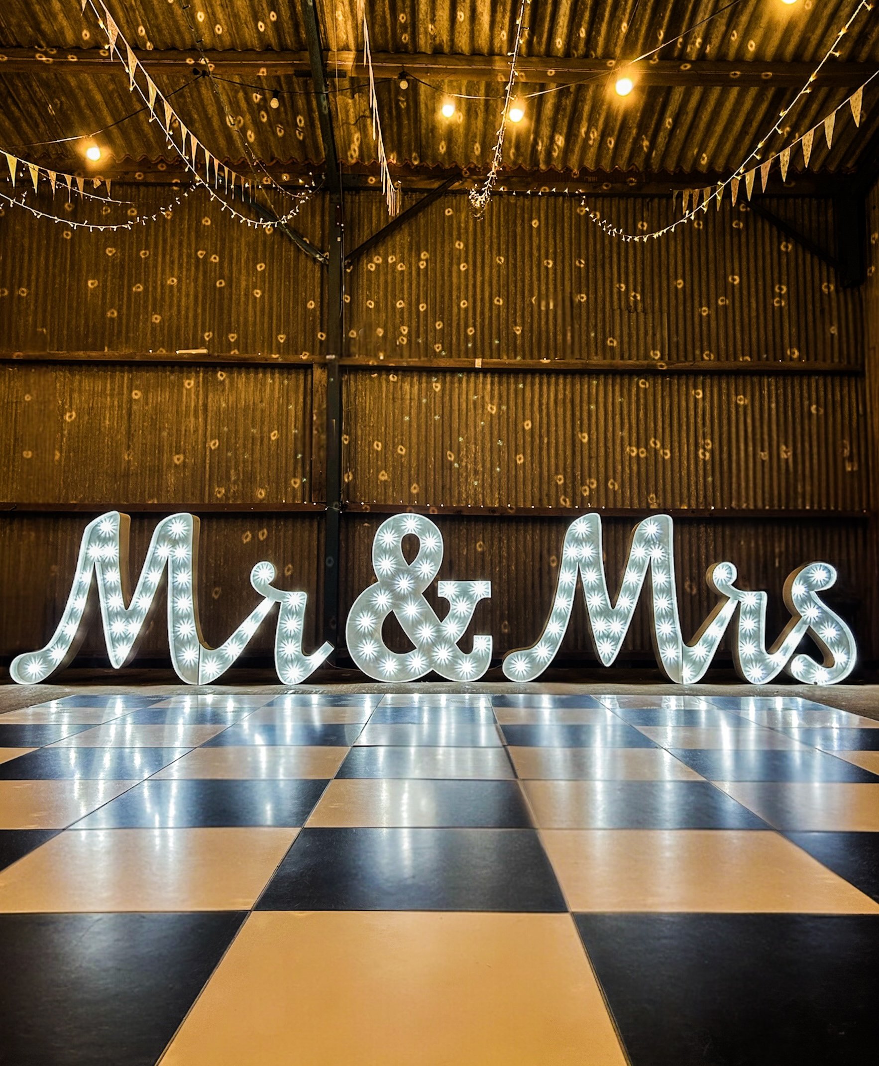 Decorative indoor space with black and yellow checkered flooring, illuminated by warm string lights, neutral chairs, and a large illuminated