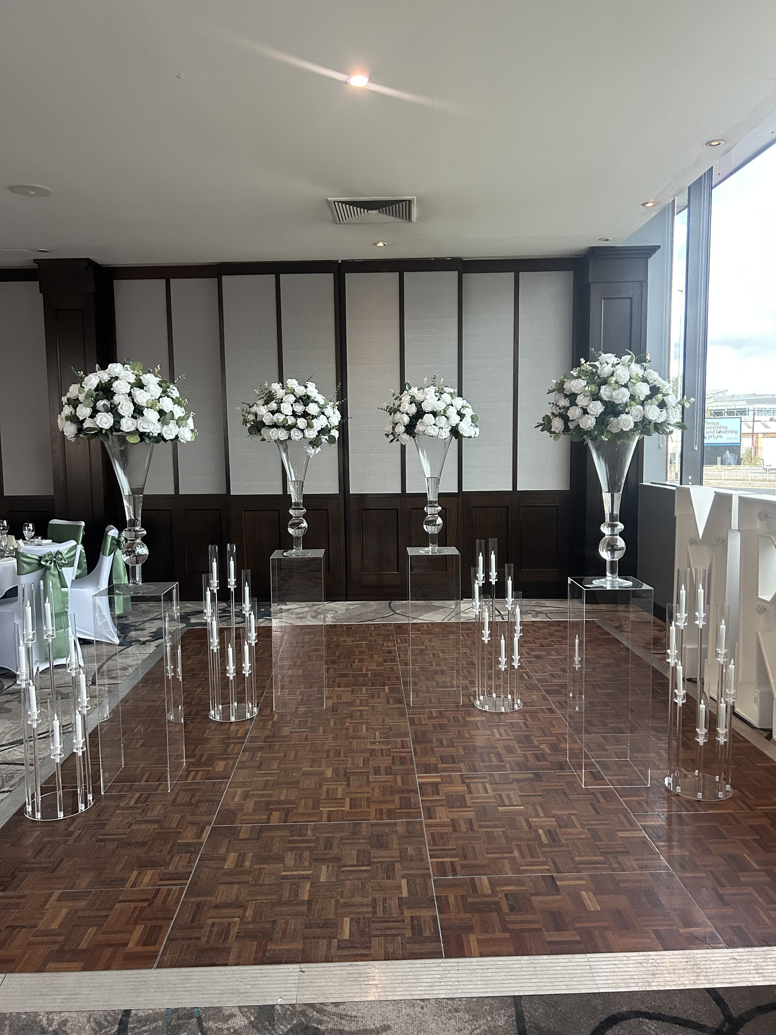 Elegant wedding or event setup with tall vases containing white roses, surrounded by clear acrylic stands with candles, in a room with wood parquet flooring and large windows. Glass vases on clear plinth columns wedding stands aisle decoration hire