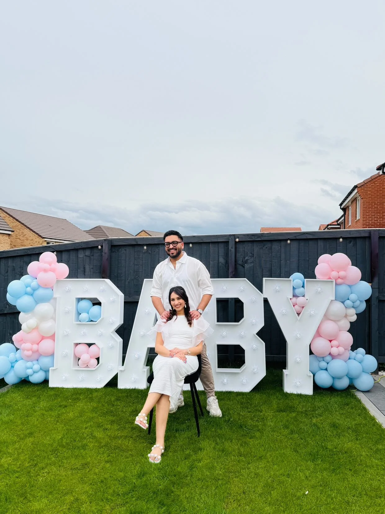 A couple celebrating a baby shower outdoors in a backyard, sitting in front of large white letters spelling 'BABY' decorated with pink, blue, and white balloons.