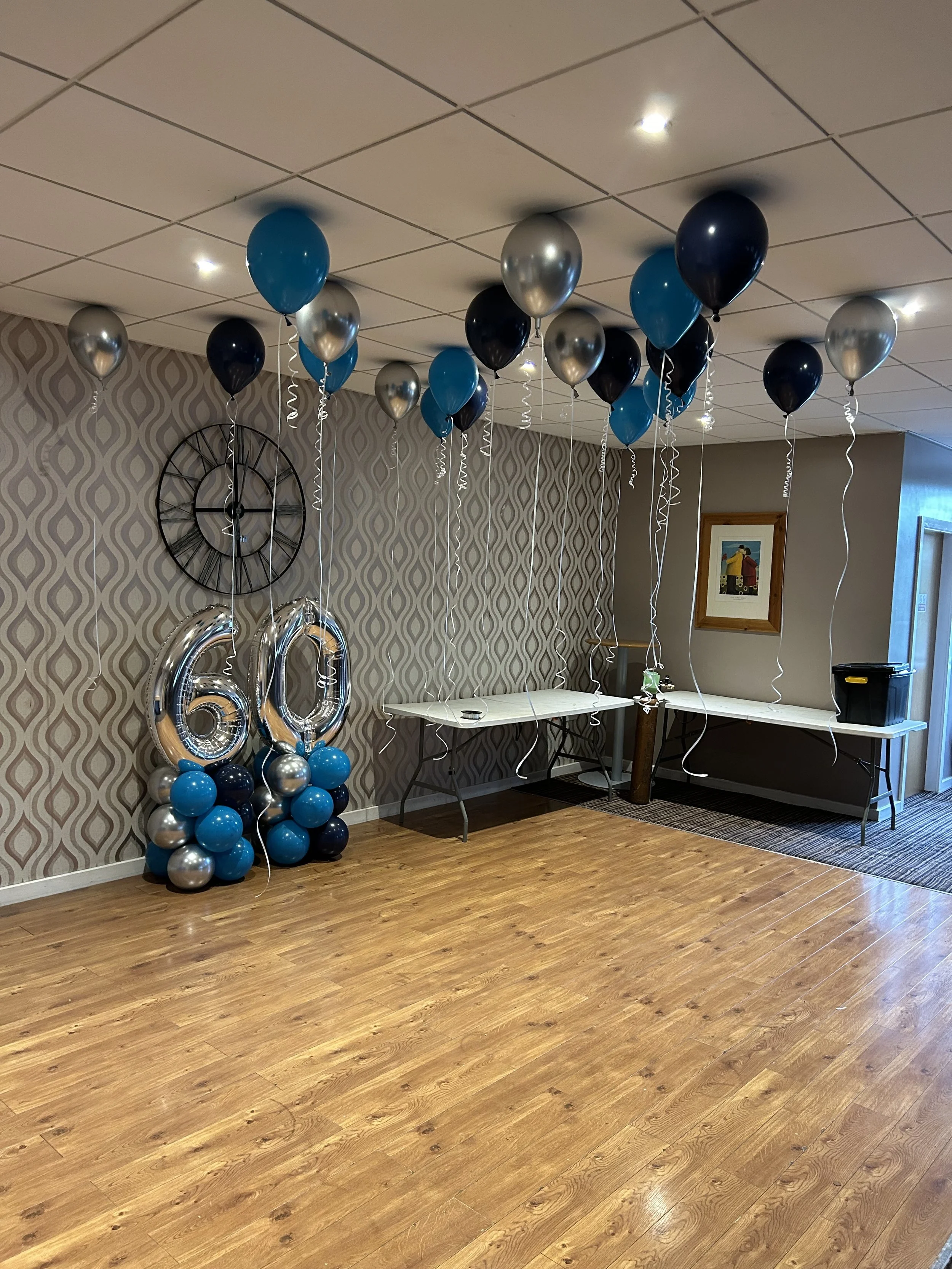 Room decorated for a 60th birthday celebration with black, blue, and silver balloons, including large silver '60' balloons, arranged in clusters on the floor. The ceiling has additional balloons tied with curly ribbons. There are two tables against t