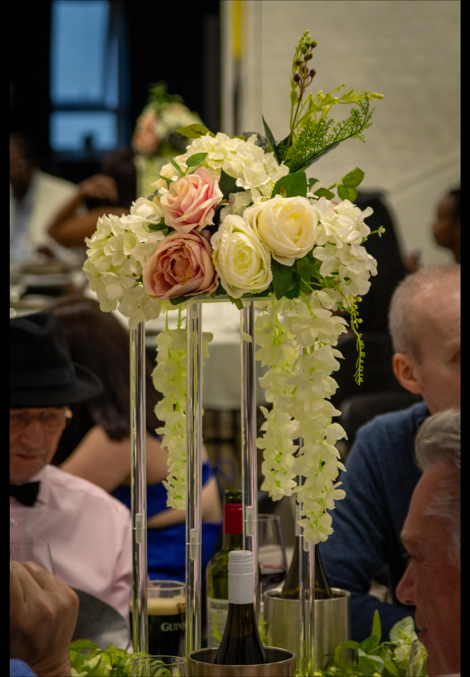 A floral centerpiece with white and pink roses and greenery on a tall stand at a formal dinner event.