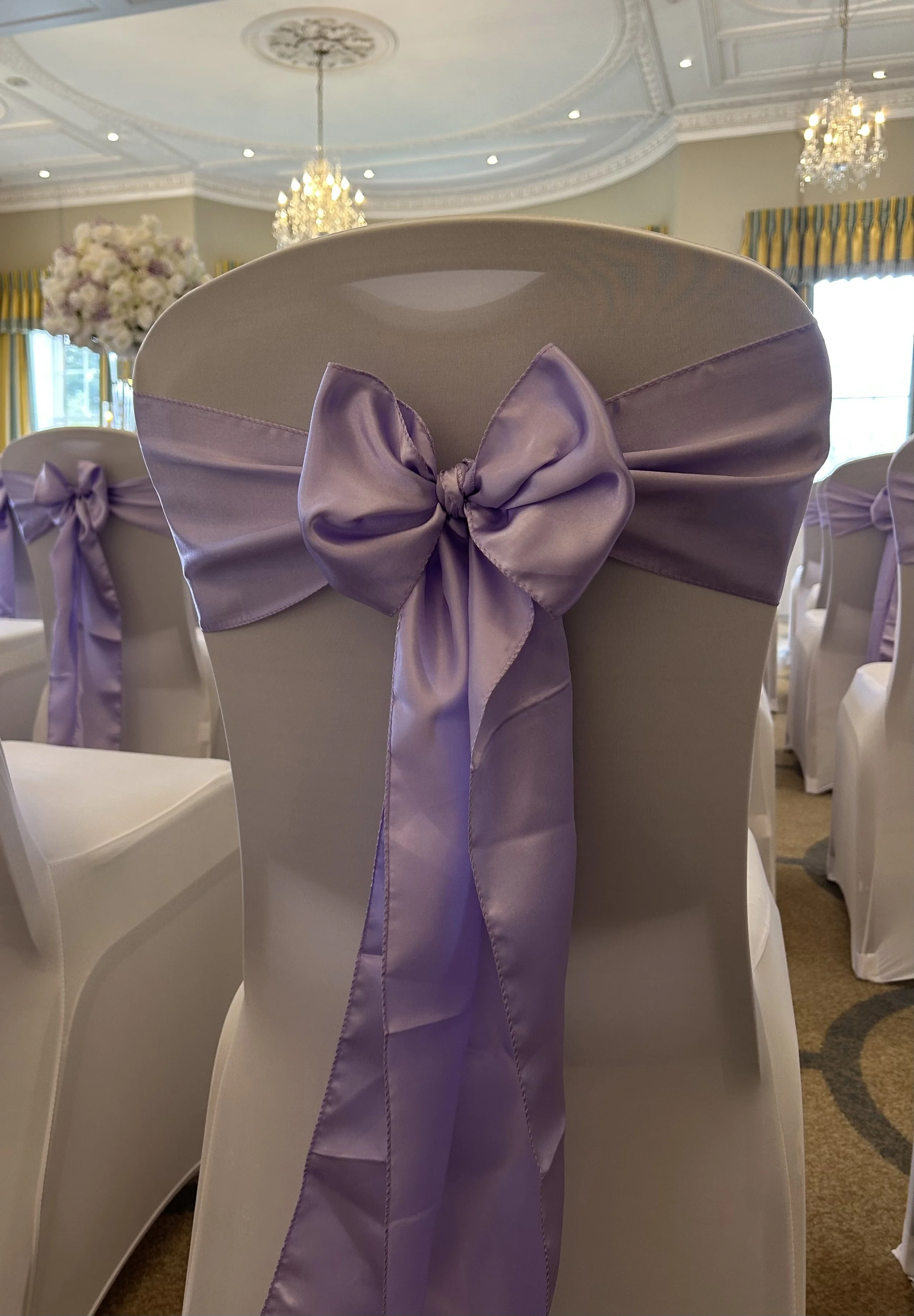 Chair with a lavender satin bow and sash, set up for a wedding or event in an elegant room with chandeliers and floral arrangements.