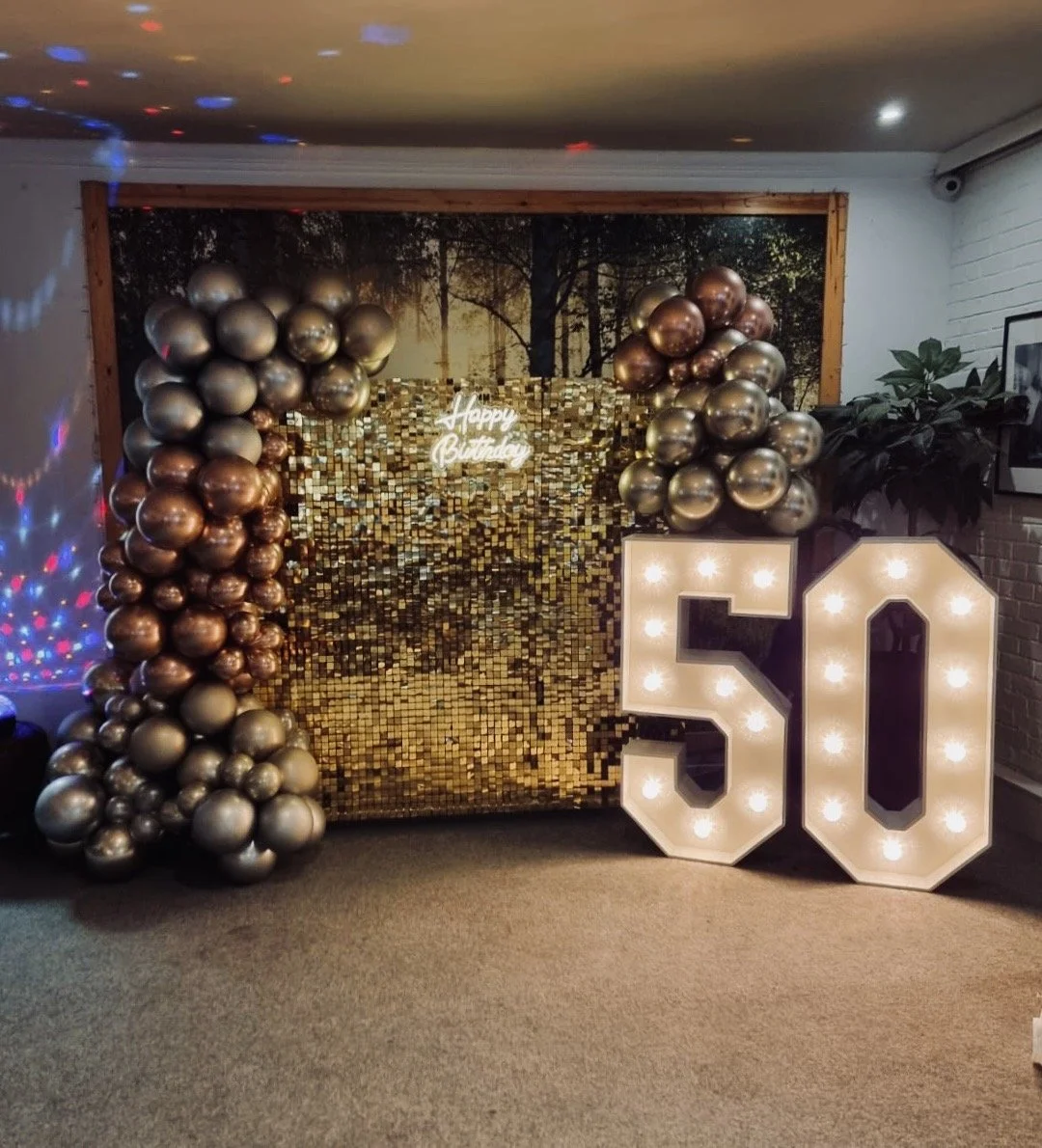 Gold and silver balloons arranged around a backdrop with a gold mosaic pattern, a lit-up marquee number '50', and a neon sign that says 'Happy Birthday' for a 50th birthday celebration.