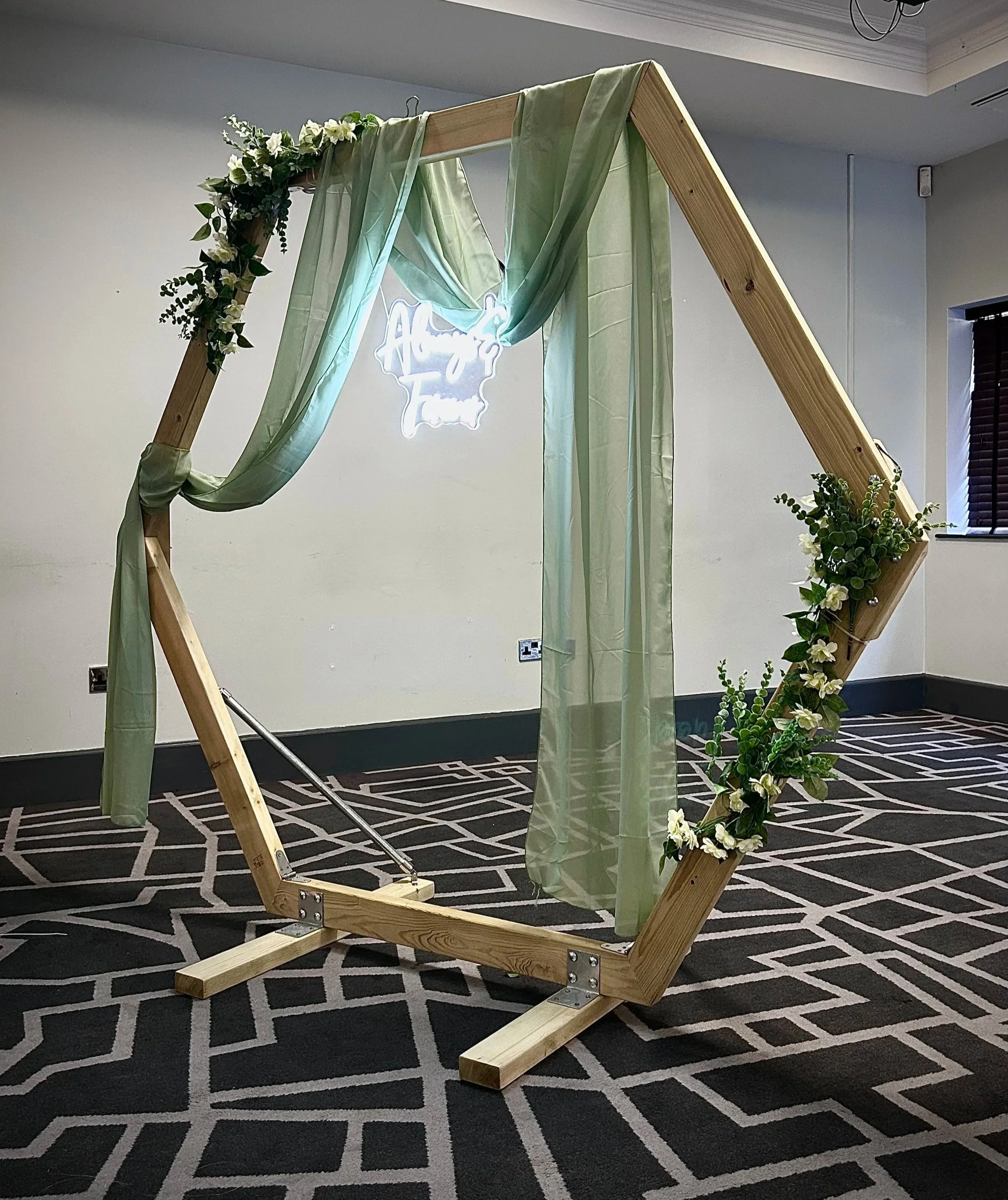 A wooden wedding arch decorated with green fabric, white flowers, and greenery, set up on a patterned carpeted floor in an indoor venue.