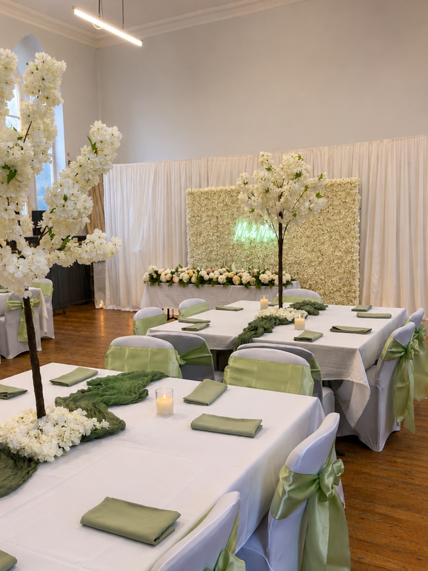 Wedding reception setup with white tables, green napkins, and white chair covers with green sashes. Centerpieces feature white flowers and green cloth accents. A decorated backdrop with a neon sign reading 'Mr & Mrs' is visible in the background.