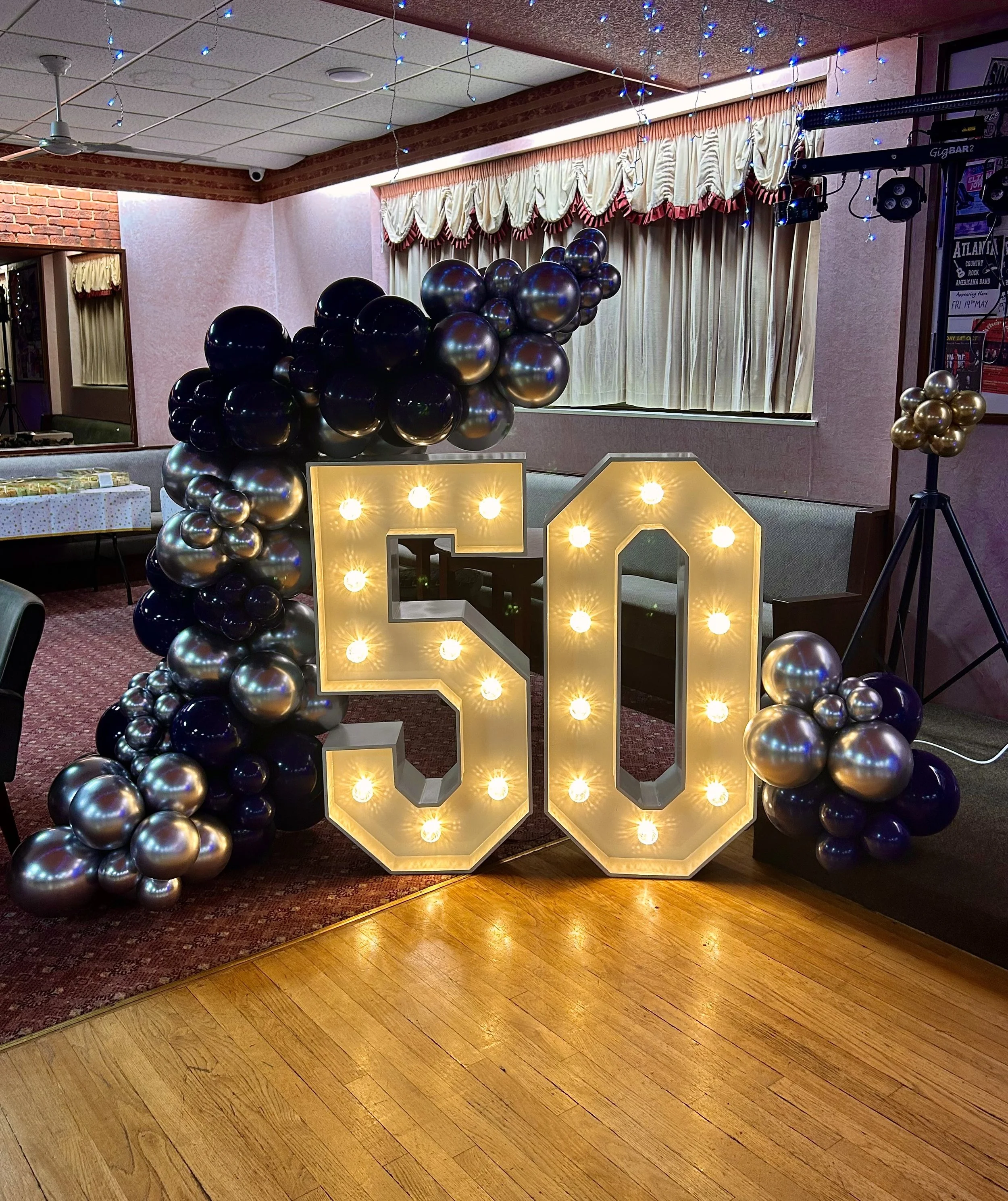 Decorative 50th birthday celebration with illuminated number 50, surrounded by black and silver balloons, inside a decorated room with curtains and string lights.