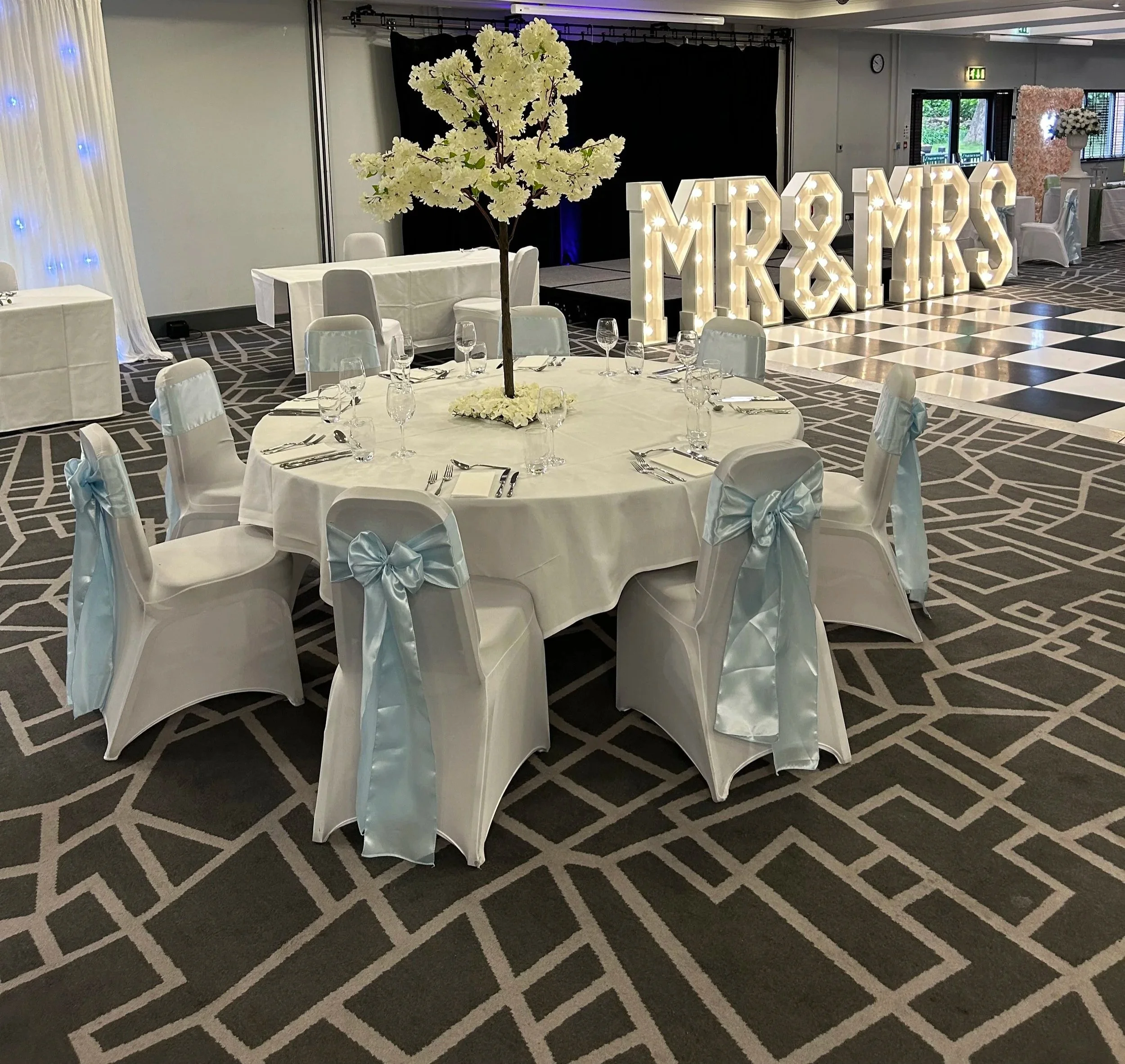 Event space decorated with white chairs with bows, a large floral centerpiece, and illuminated letters spelling 'MR & MRS' in the background, with tables set with glasses and utensils, and a checkered floor pattern.