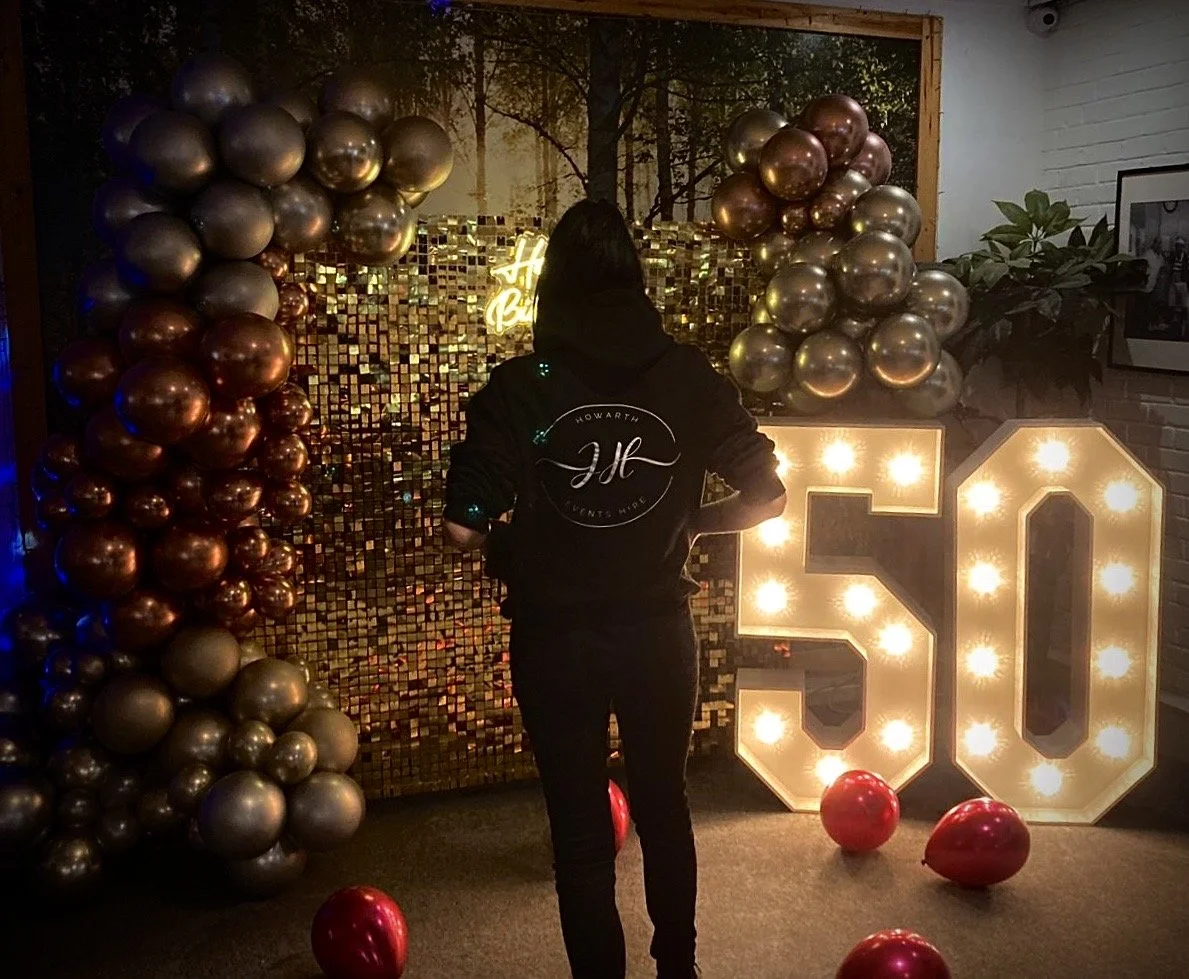 Person standing in front of a decorated backdrop with gold balloons, illuminated large number 50 sign, and red balloons on the floor, celebrating a 50th birthday.
