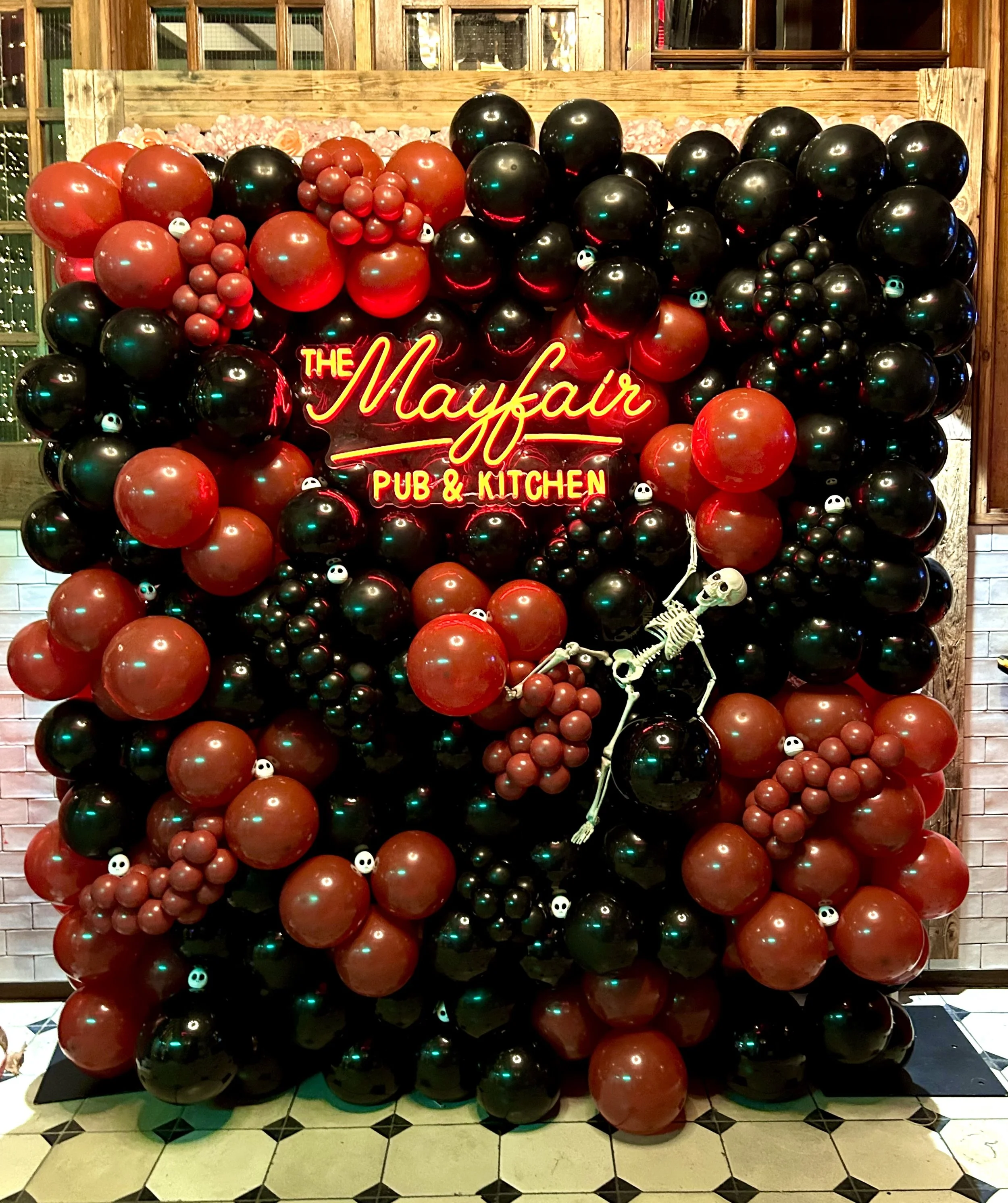 Decorative display with black and red balloons, a neon sign reading "The Mayfair Pub & Kitchen," small skull and skeleton props, and a background of brick and wood walls.