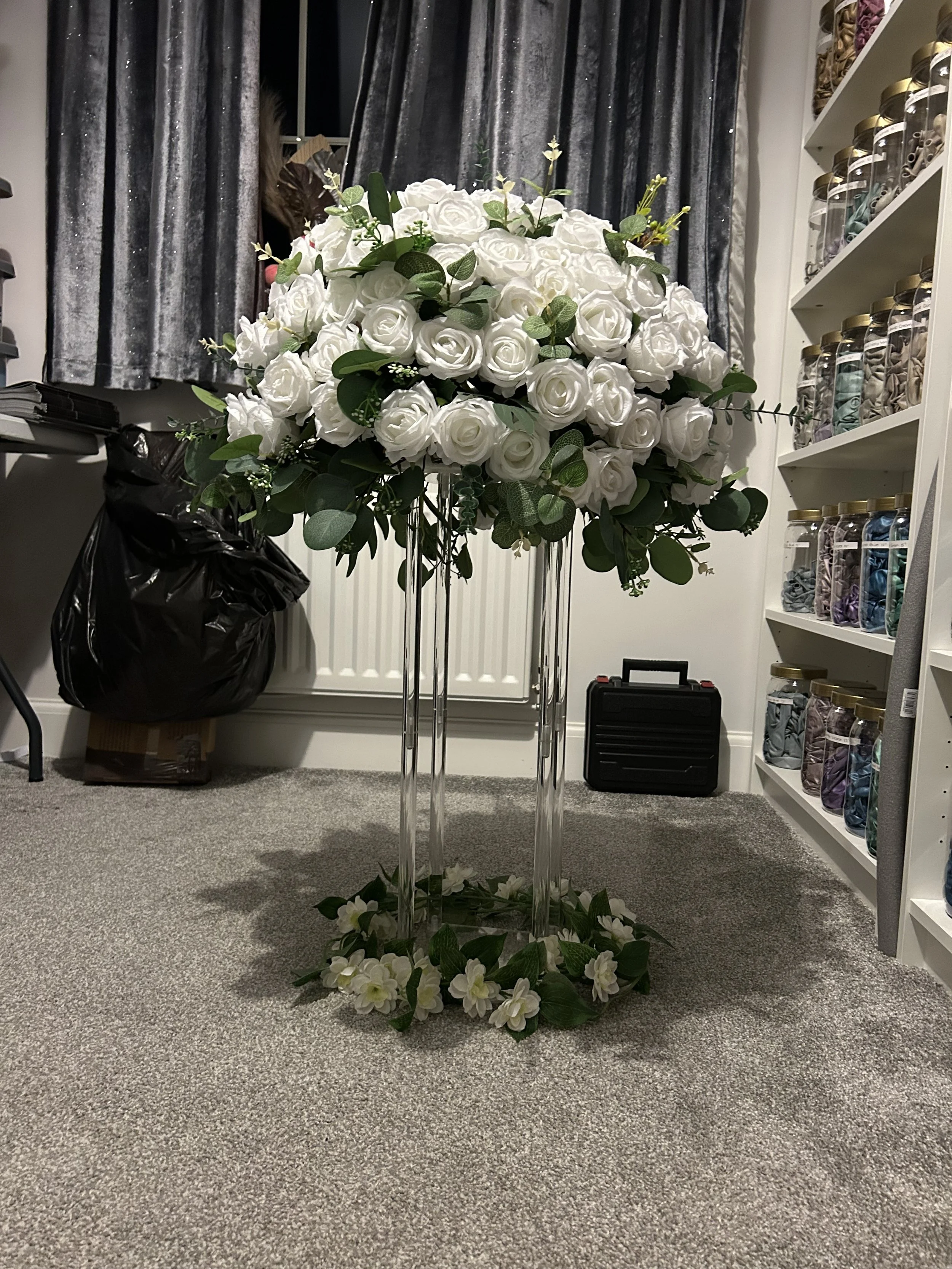 A tall floral arrangement of white roses and green leaves, supported by clear acrylic stands, on a gray carpeted floor in a room with shelves of jars and blue curtains in the background.