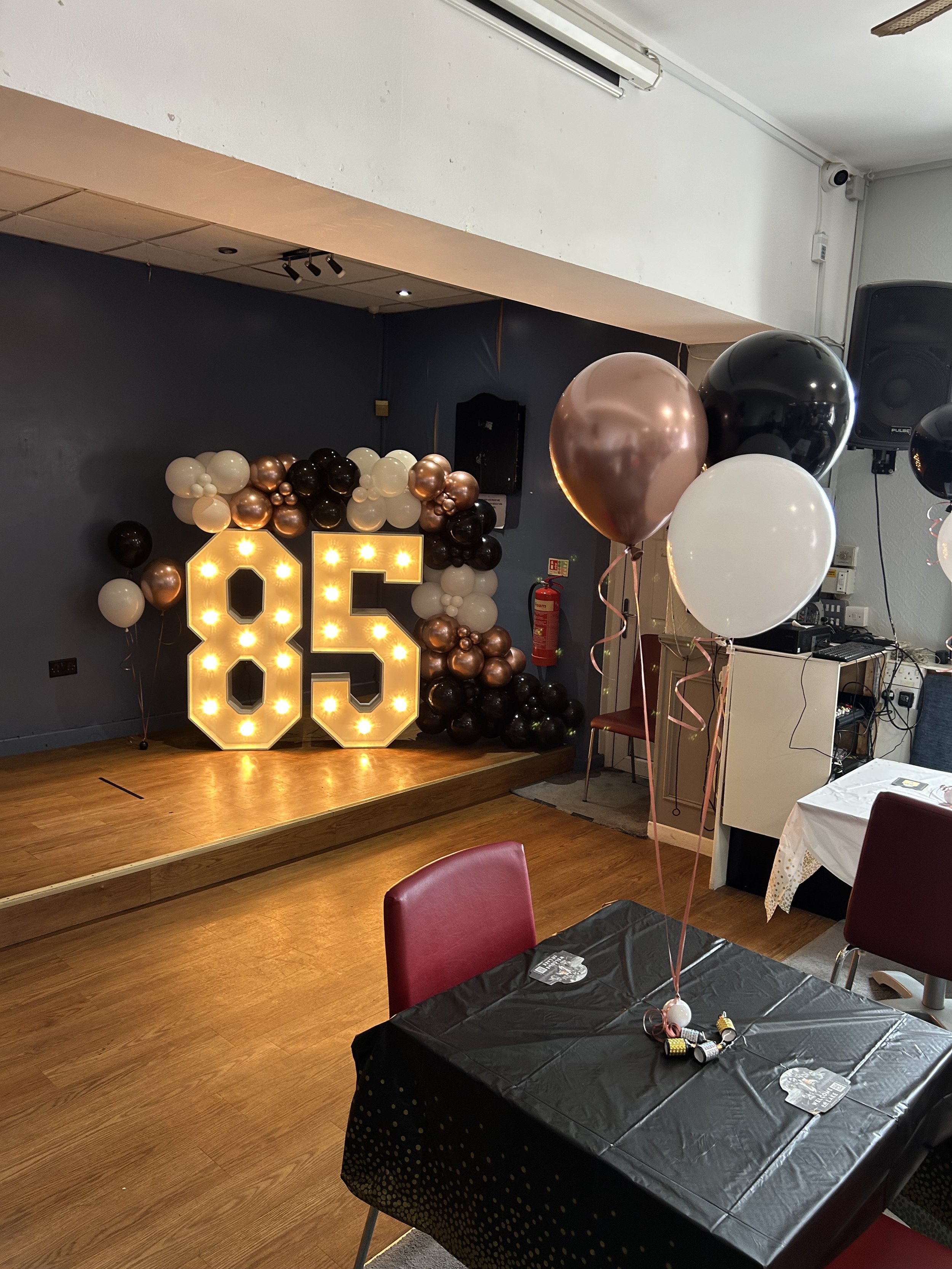 Decorated party venue with balloons, illuminated number '85', and tables with black tablecloths.