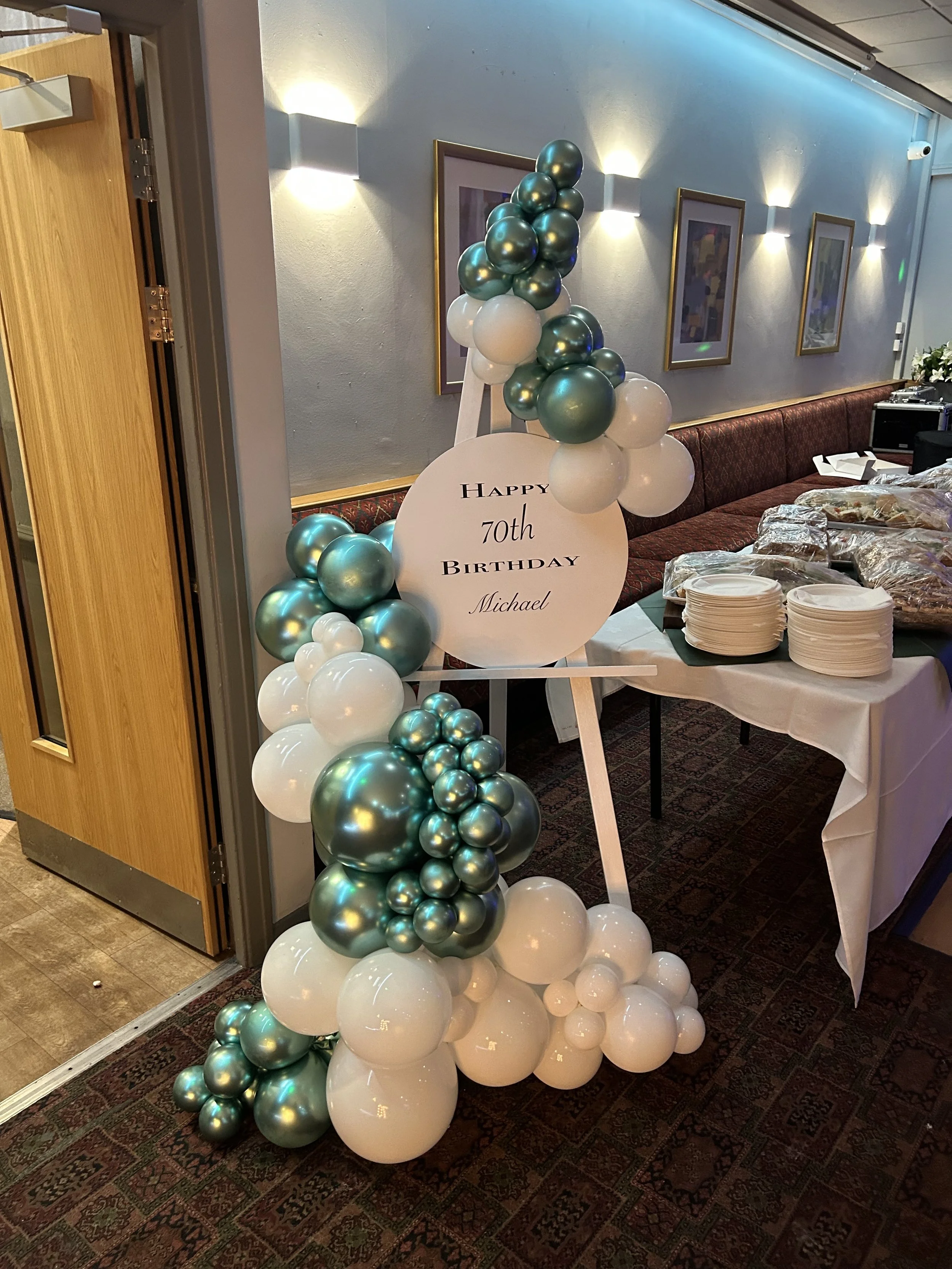 Celebratory balloon arrangement with green and white balloons surrounding a sign reading "Happy 70th Birthday Michael" at a decorated party venue with plates and food on a table nearby.