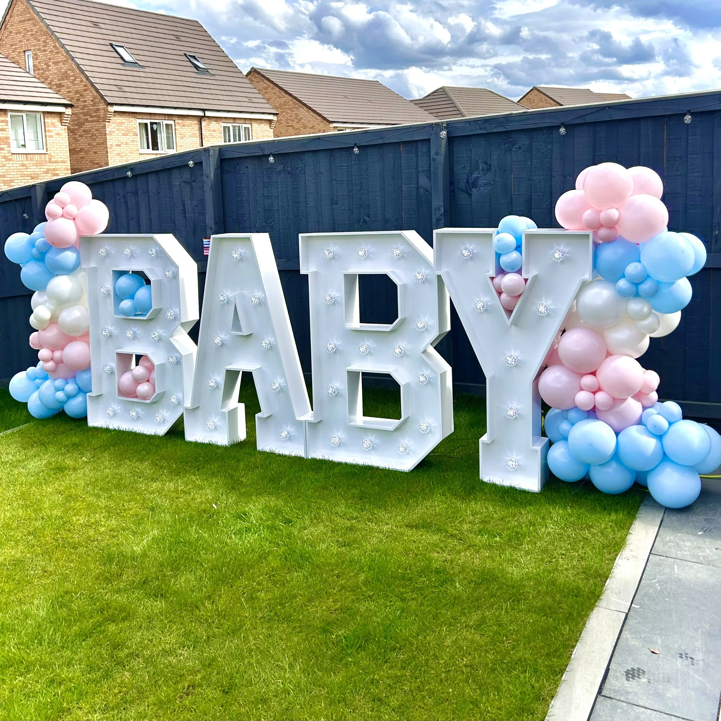 Large illuminated letters spelling 'BABY' with pink, blue, and white balloons on each side, set on green grass with a blue fence and houses in the background.