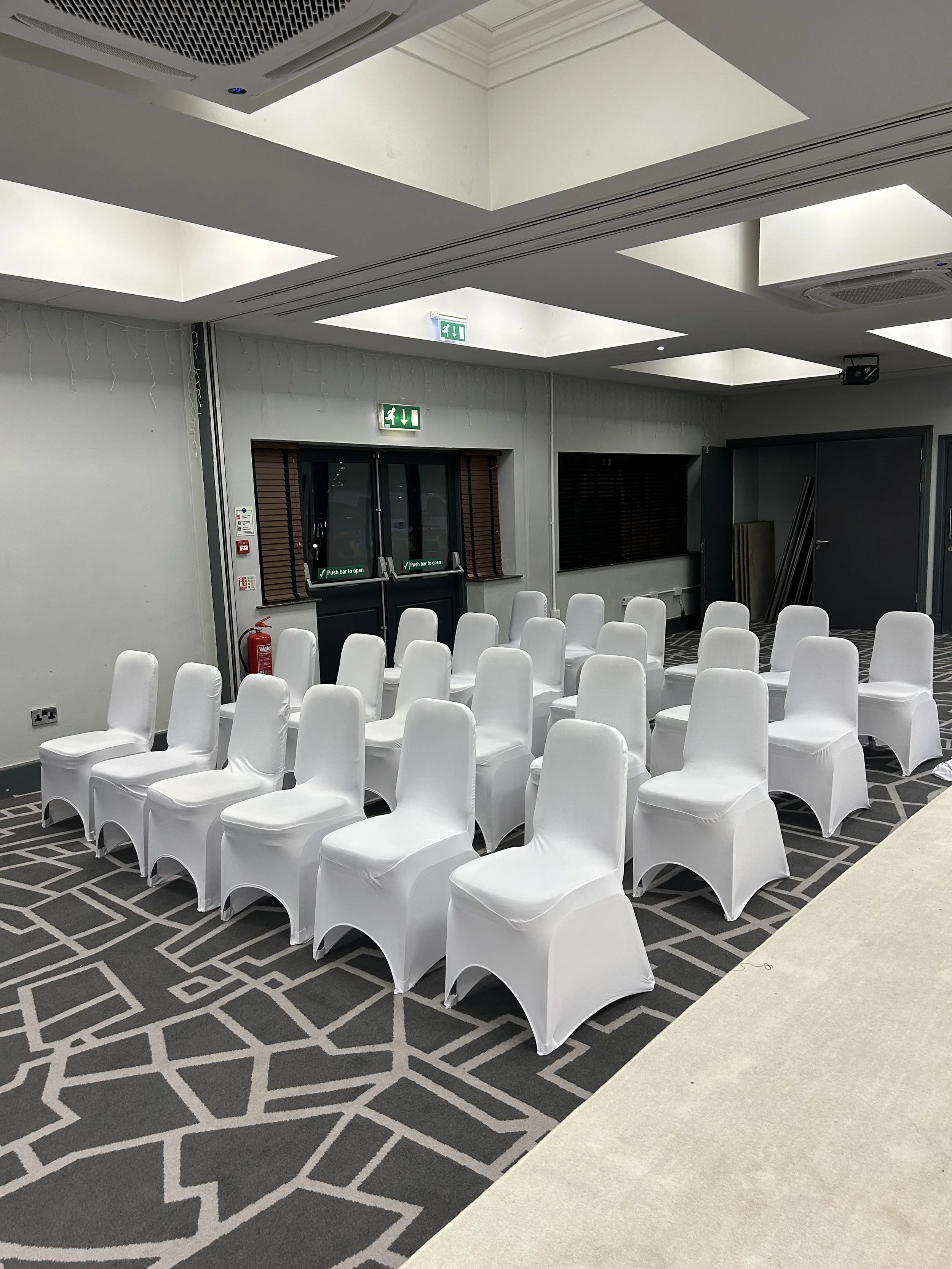 Several rows of white chairs arranged in a conference or event room with gray patterned carpet, grey walls, and black window blinds. There are emergency exit signs above two doors and a smoke alarm on the wall. Ceiling has square light panels and air vents.
