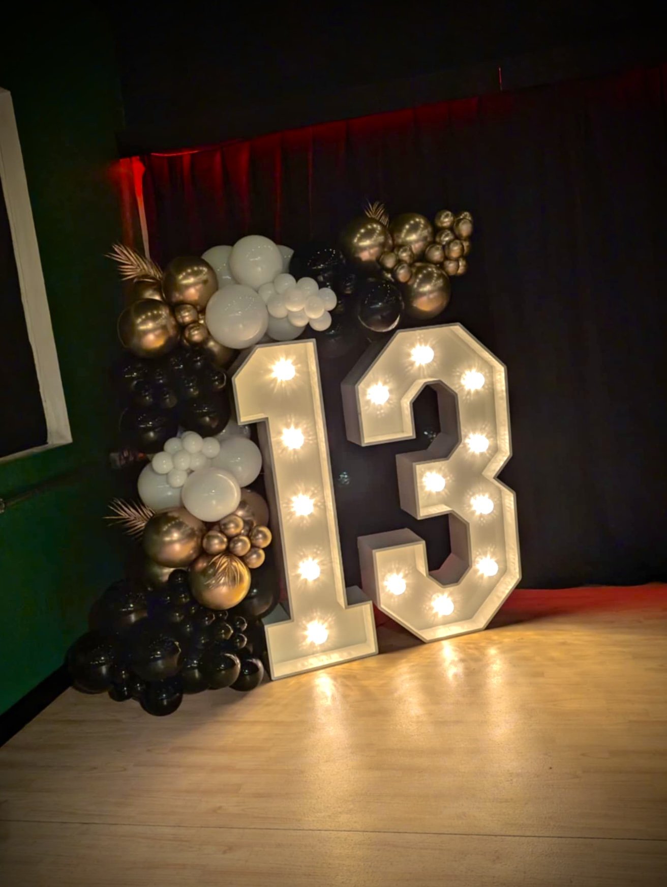 Decorative birthday display with large illuminated number 13 and black, gold, white, and metallic balloons arranged on a wall.