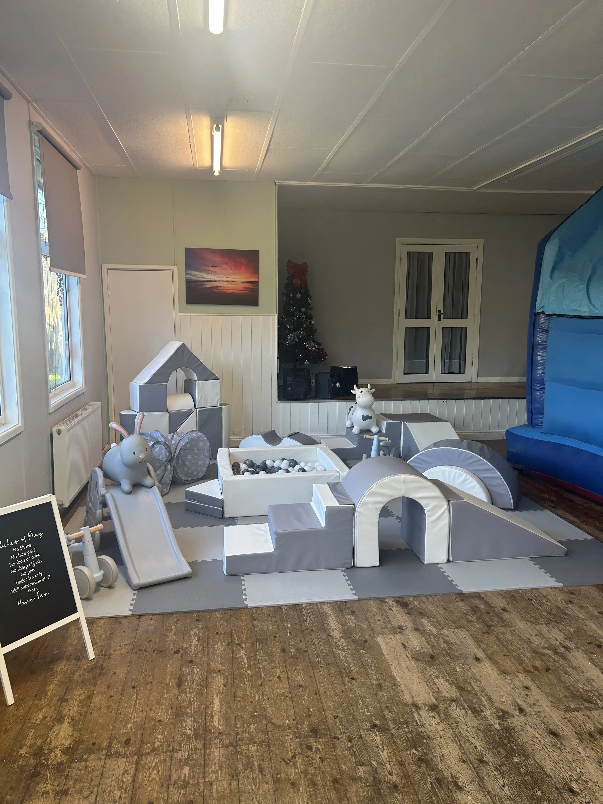 Indoor children's play area with soft padded structures, slide, ball pit, and plush animal toys, in a room with wooden floor, windows, and a decorated Christmas tree in the background.