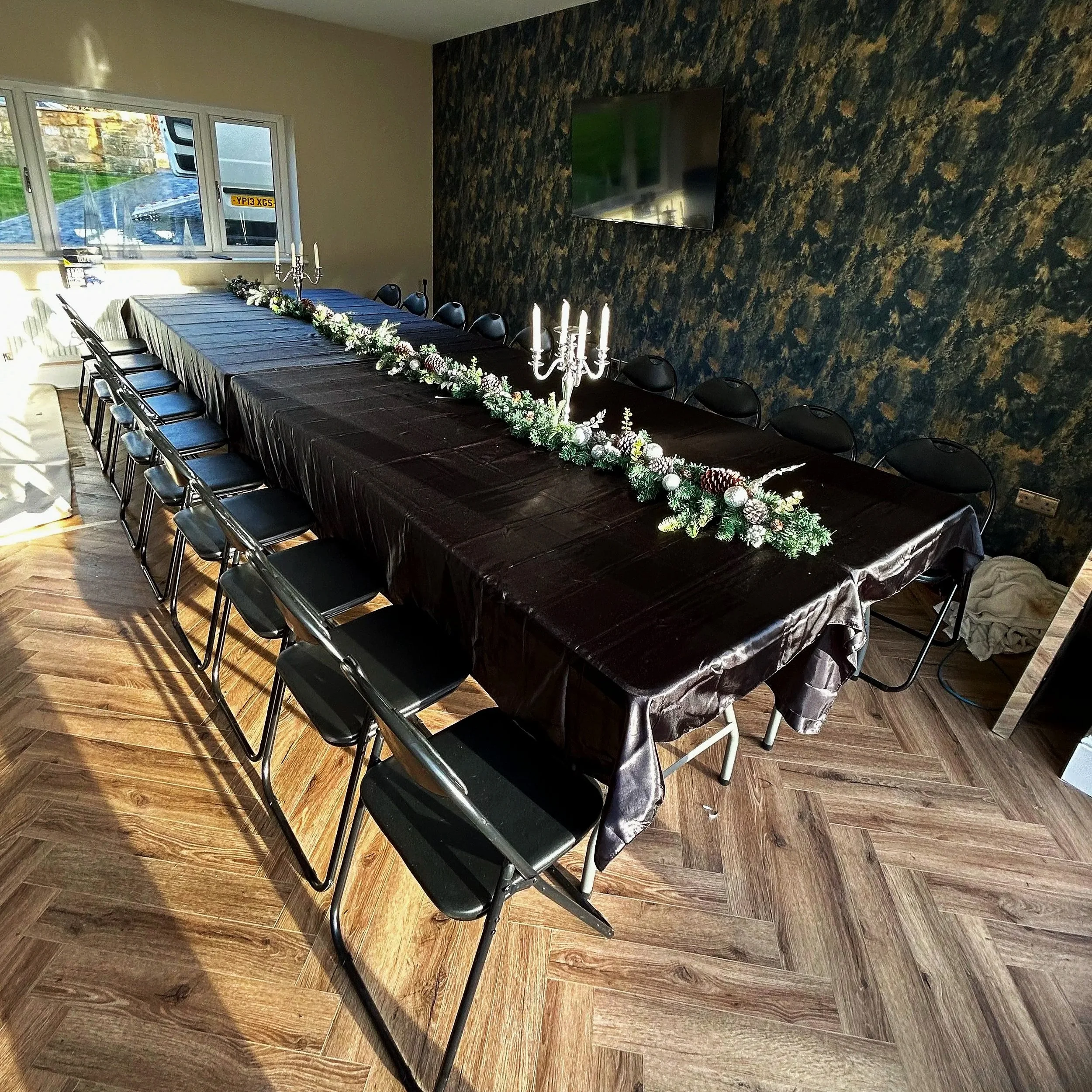 A long dining table covered with a dark tablecloth, decorated with a greenery and berry garland, and topped with candelabras with white candles, set in a room with wooden flooring, a dark feature wall, and a flat-screen TV on the wall.