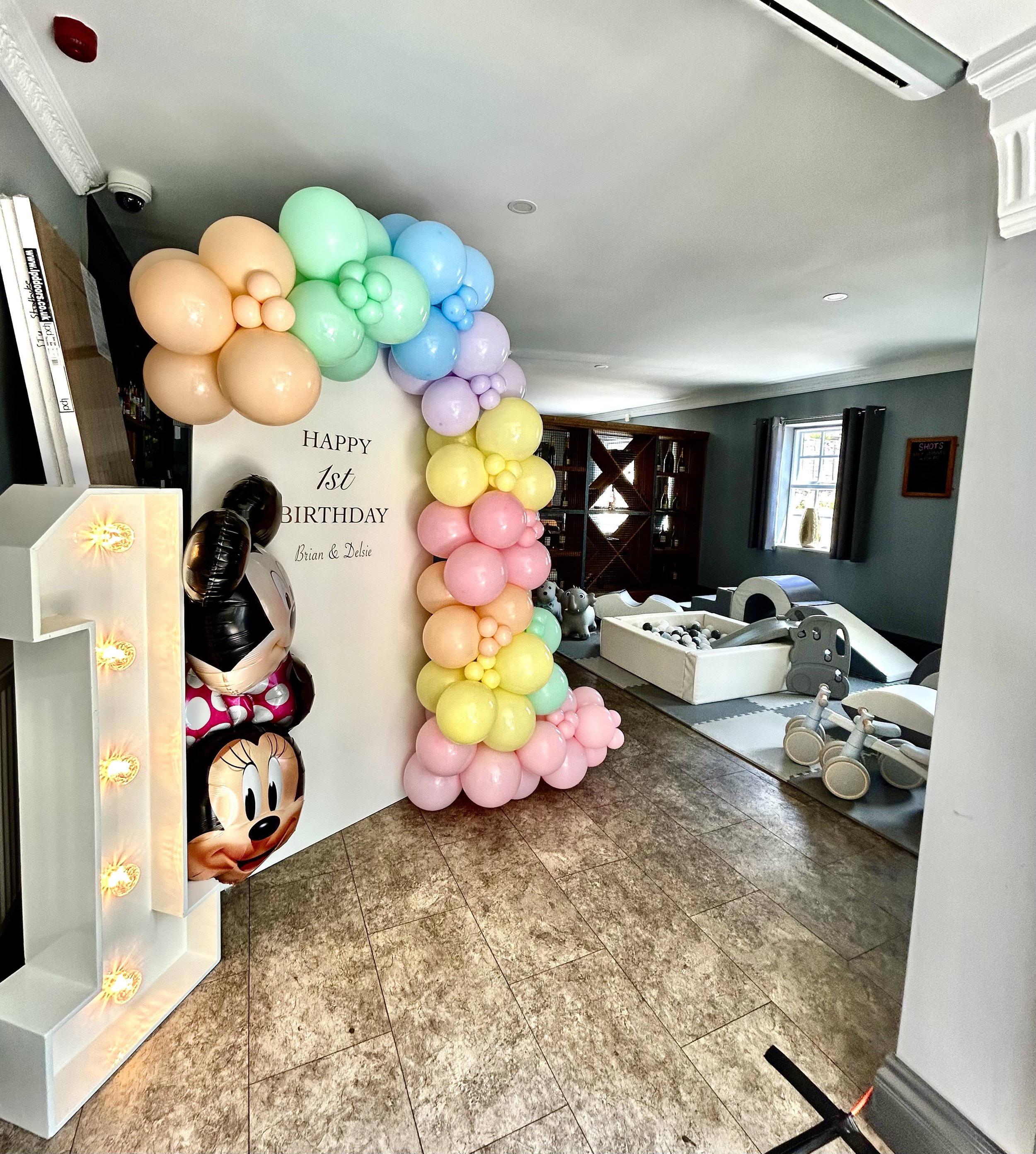 Decorated indoor space for a first birthday celebration featuring an arch of pastel balloons in yellow, pink, green, blue, and purple, a Minnie Mouse themed balloon, and a sign that reads "Happy 1st Birthday" with names Brian and Delsie. The room includes a foosball table, chairs, and a window with blue curtains.