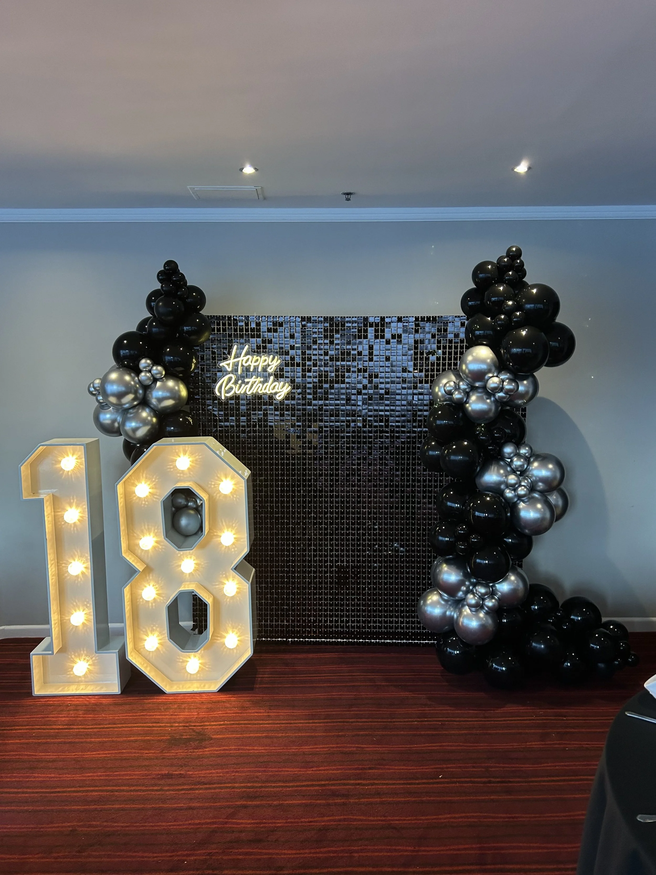 Decorative birthday display with large illuminated number 18, black and silver balloons, a black tiled background, and a lit sign saying 'Happy Birthday'.