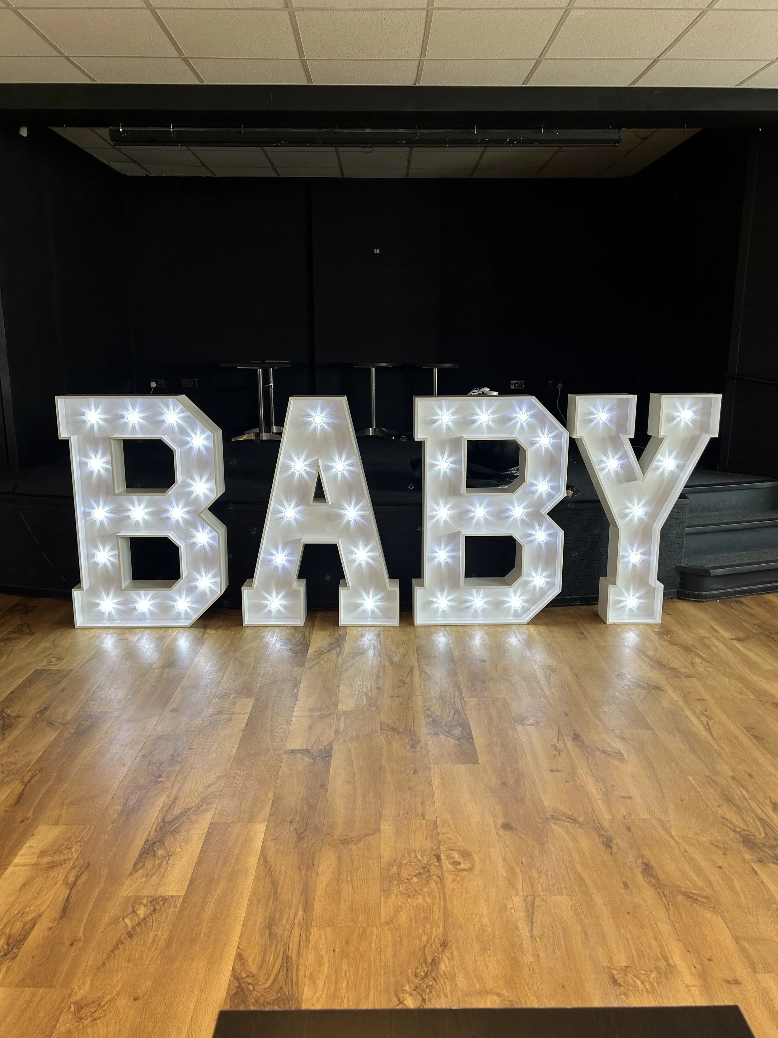 Large illuminated letters spelling 'BABY' are positioned on a wooden floor against a black background in a dimly lit room.