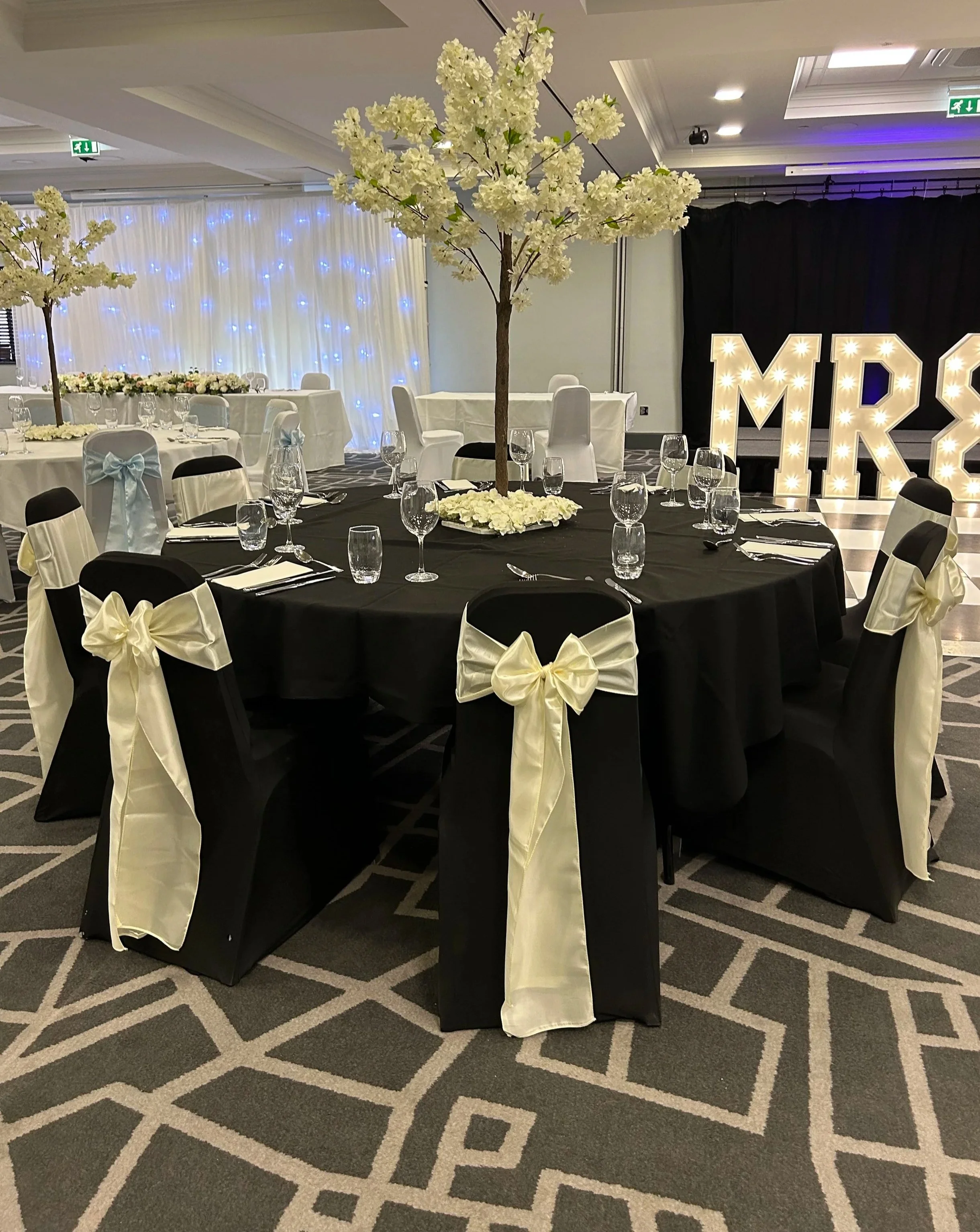 Decorated banquet hall with round tables, white chair covers with bows, tall artificial white cherry blossom trees, and large illuminated letters spelling 'MRS' in the background.