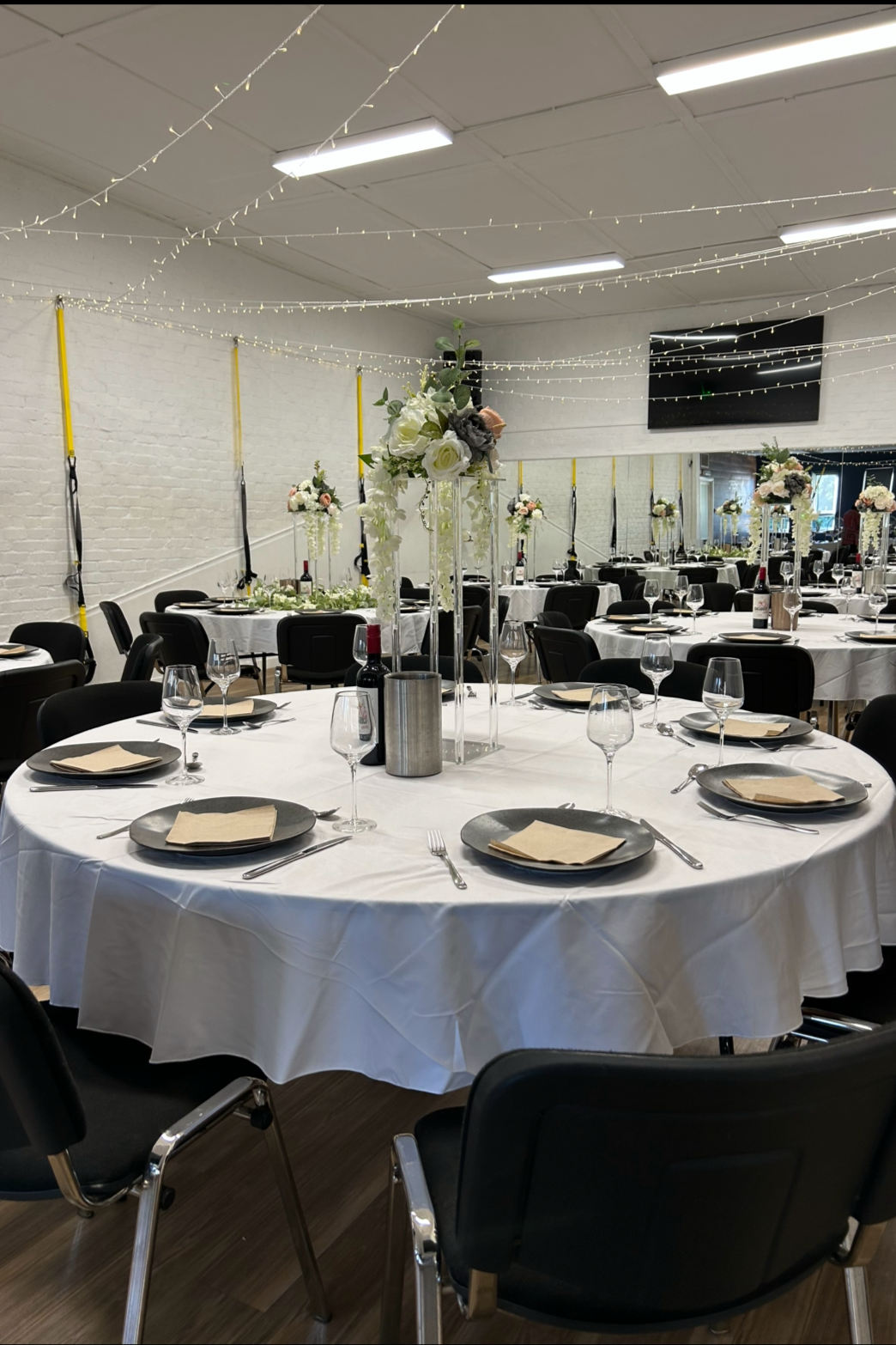 Decorated banquet hall with round tables covered in white tablecloths, each set with black plates, beige napkins, wine glasses, and cutlery. Tall floral centerpieces with white and blush flowers and greenery dominate the tables. String lights hang from the ceiling, creating a festive atmosphere.