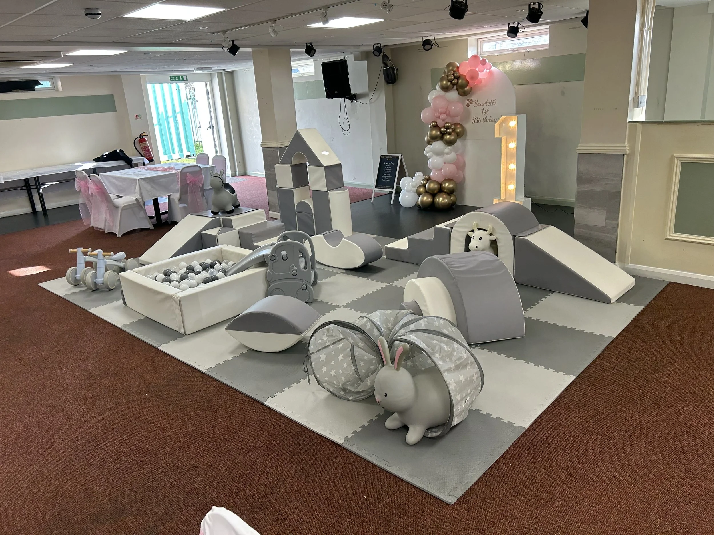 Indoor kids' play area with soft gray and white foam climbing structures, a ball pit, a rabbit-shaped tunnel, and various toys. In the background, there are decorated chairs, a table, and a birthday decoration with balloons, a large number one, and a