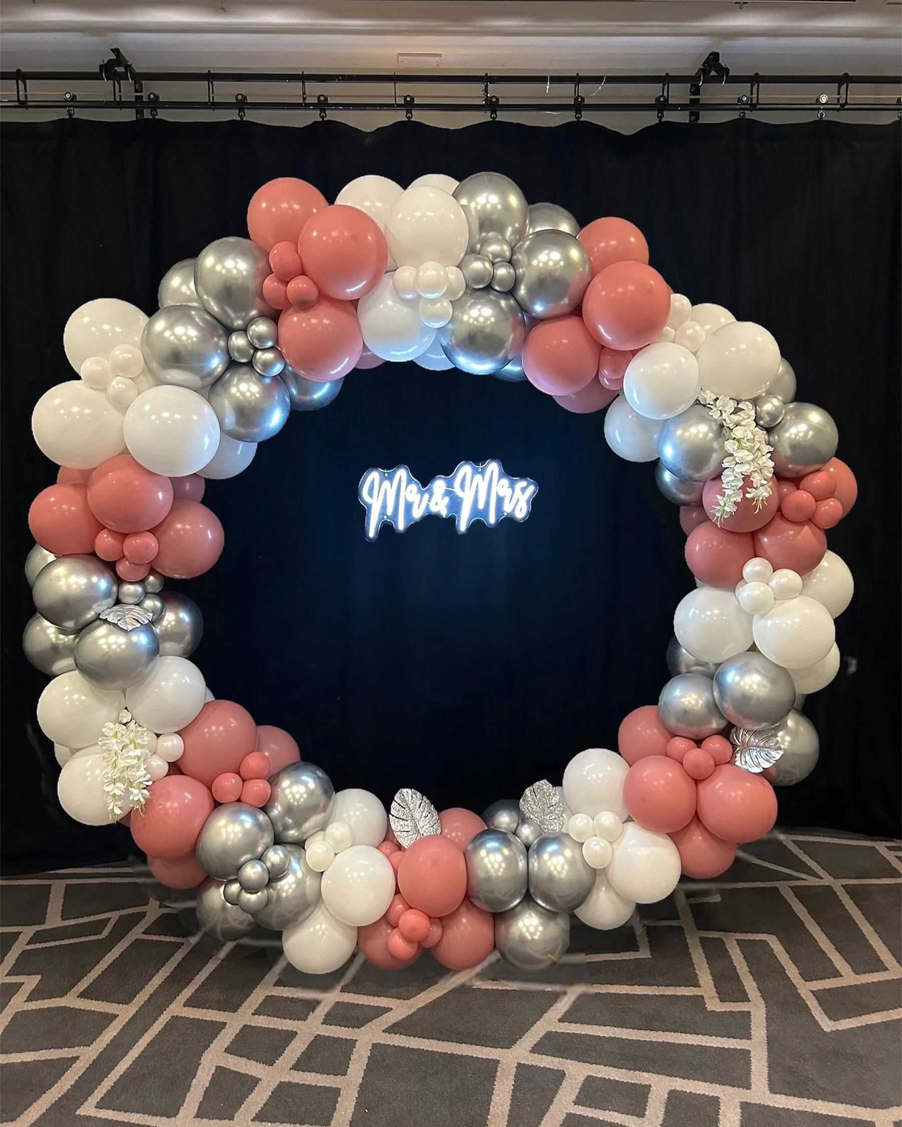 A circular balloon arrangement with pink, white, and silver balloons, decorated with white flowers and metallic leaves, with a sign in the center that reads 'Mr & Mrs' against a black background.